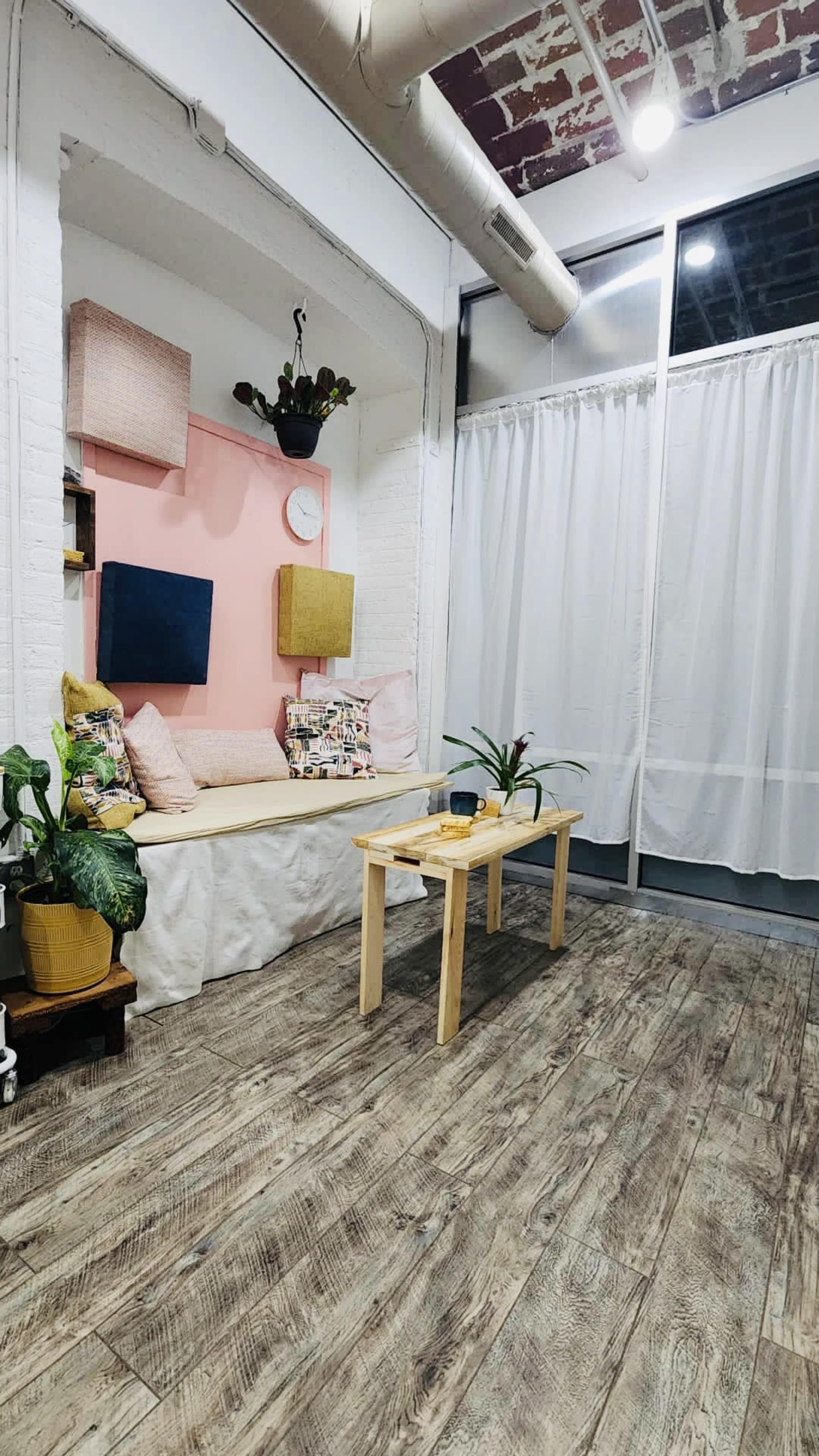 A cozy room with a seating nook against a pink wall, a small wooden table in the center, and plants in the corners.