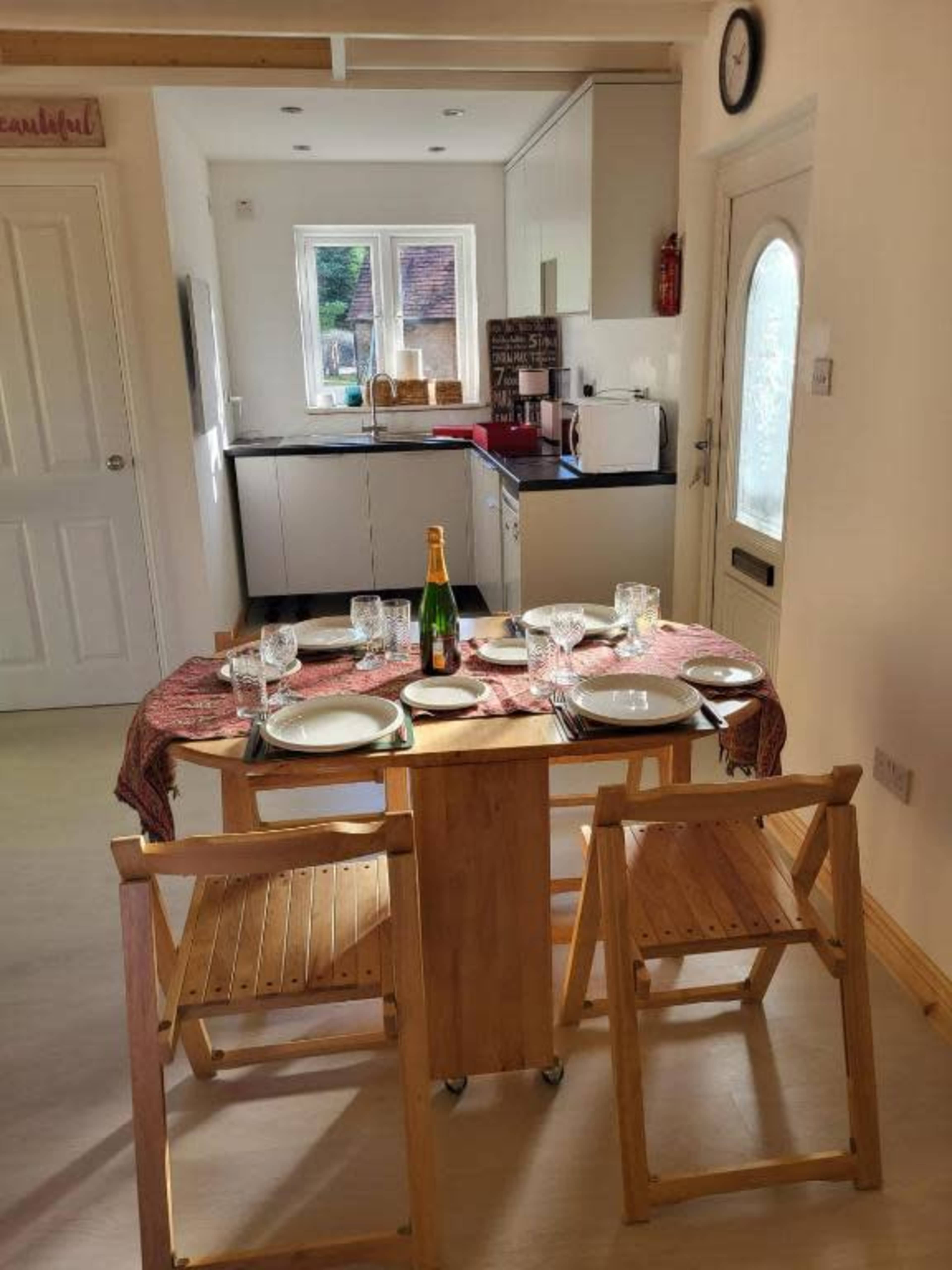A round table set with plates and glasses is positioned in a kitchen area with cabinets and appliances in the background.