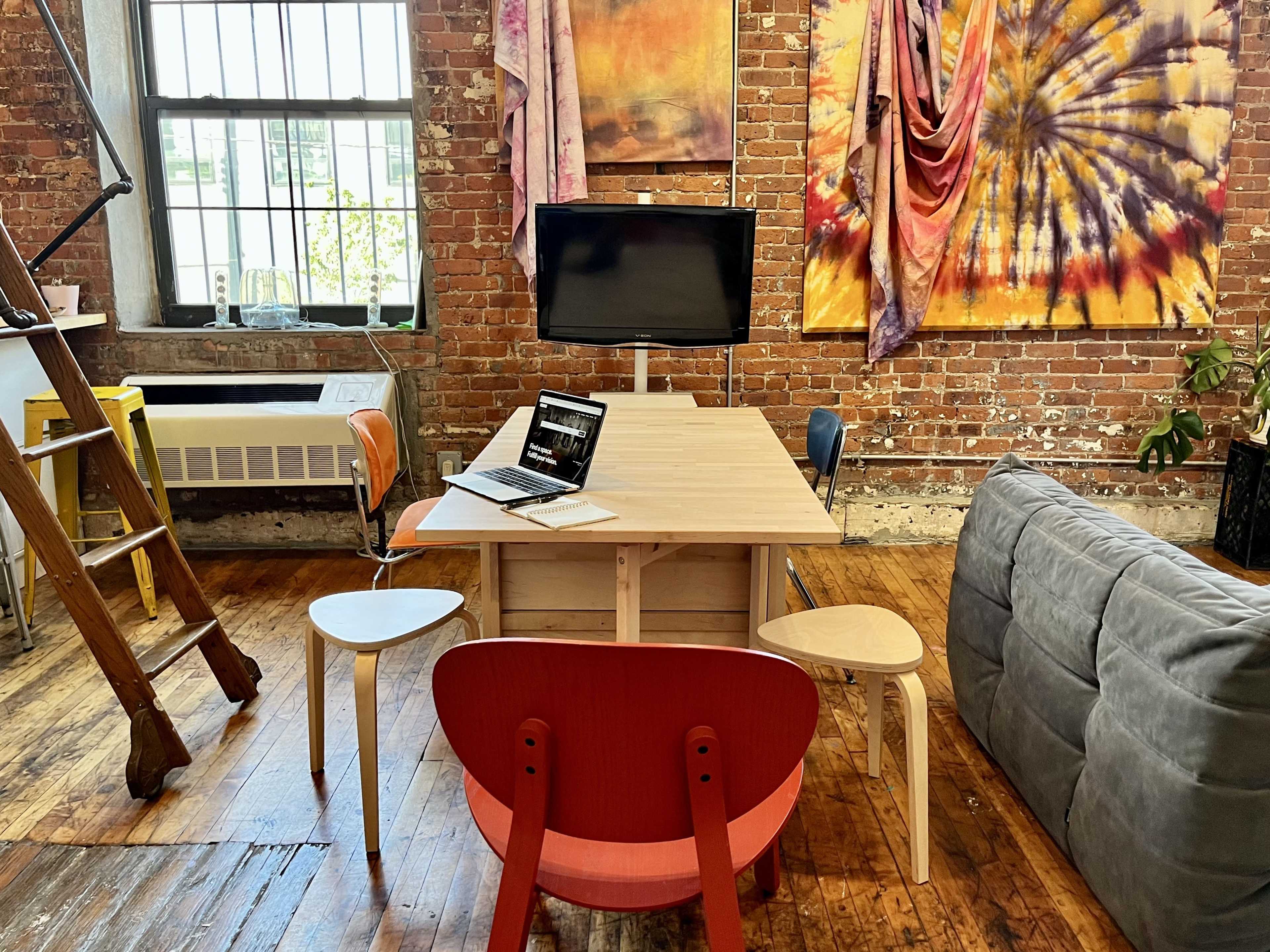 A wooden table surrounded by colorful chairs and a sofa is set up in a spacious room with exposed brick walls and art pieces hanging nearby.