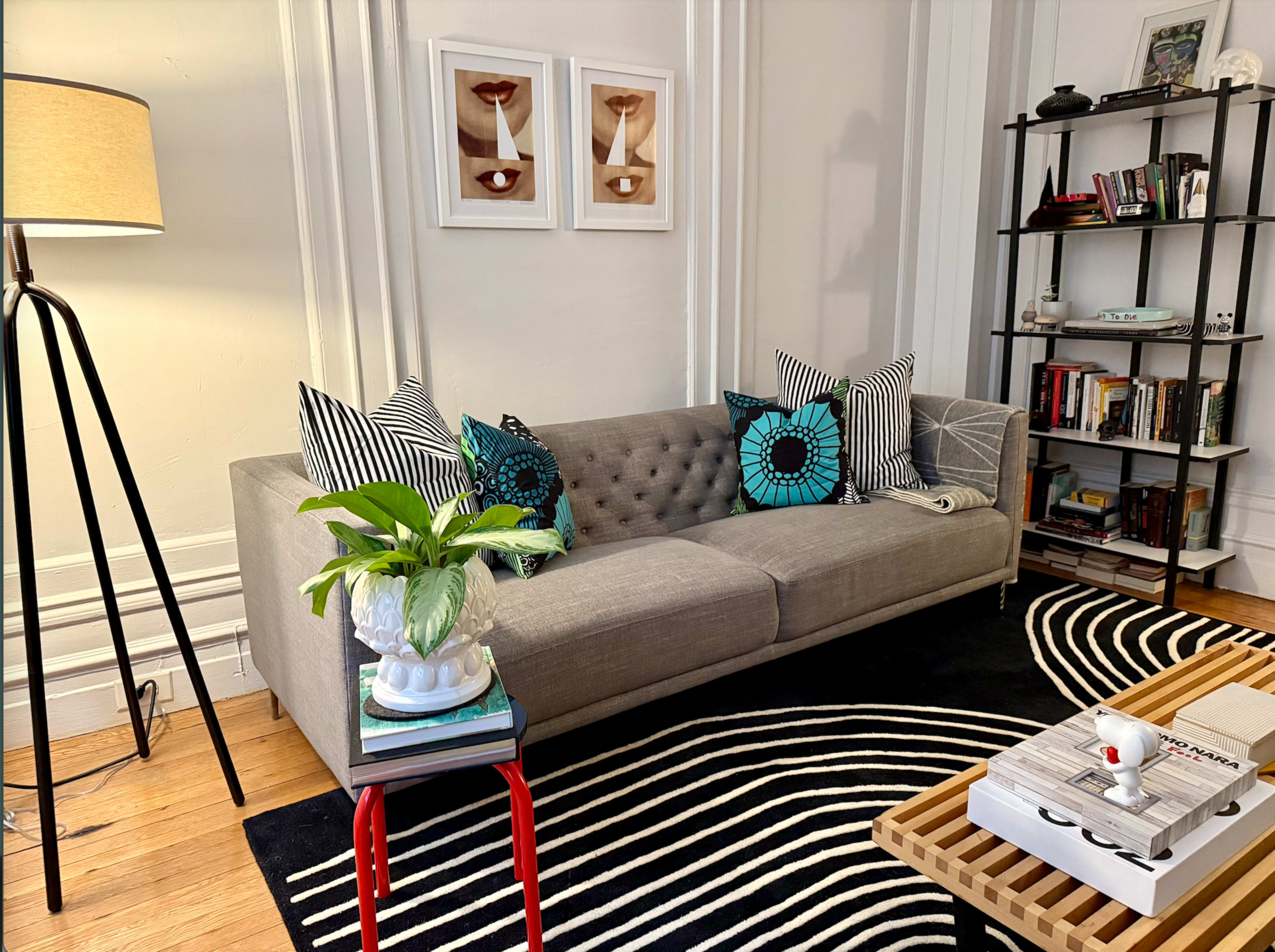 The image shows a modern living room featuring a gray sofa with patterned cushions, a lamp, a bookshelf, and a coffee table on a striped rug.