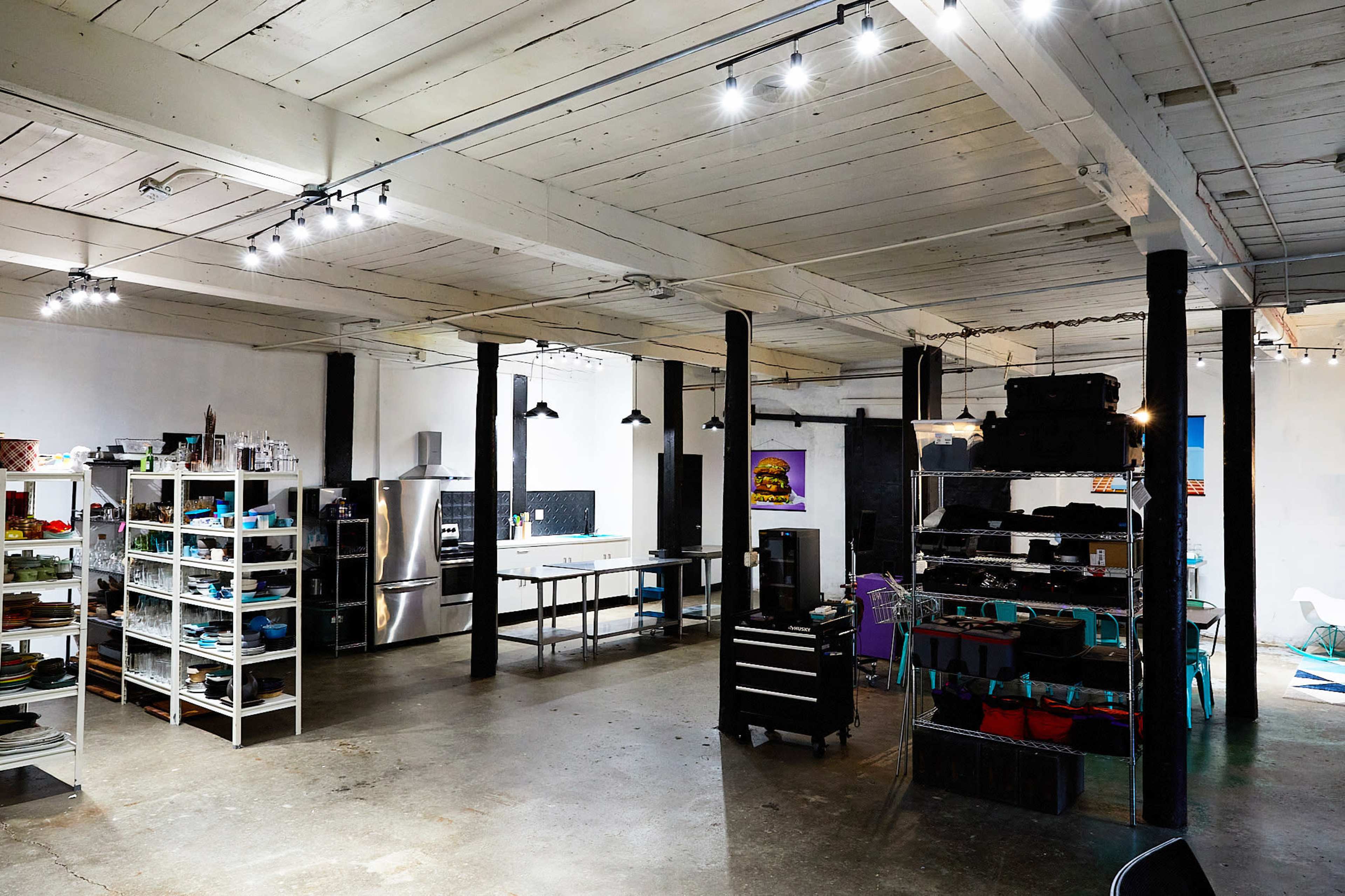The image shows a spacious kitchen and workspace with white walls, metal appliances, shelves filled with kitchenware, and various tools arranged on tables.