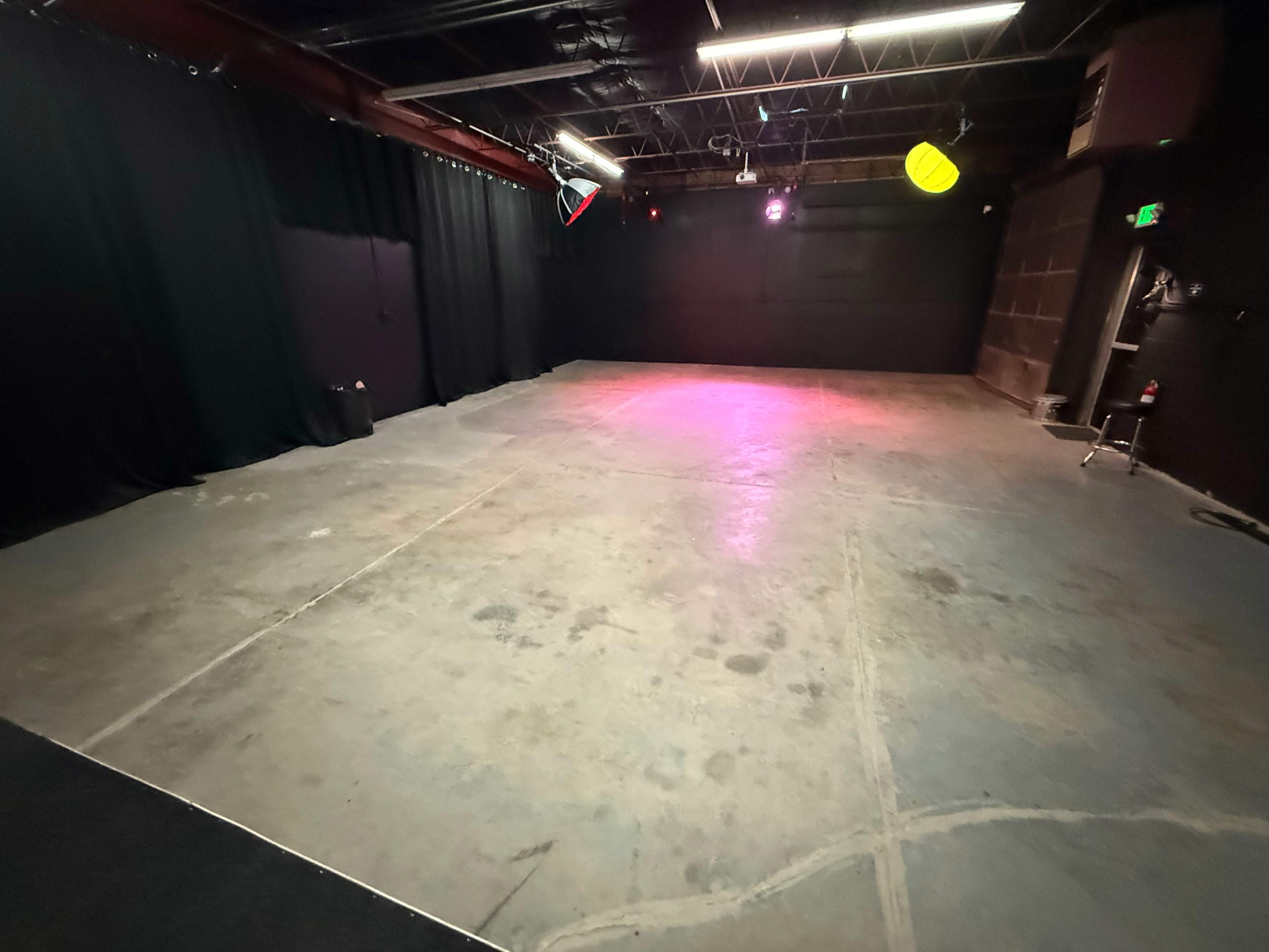 The image shows a large, empty performance space with a concrete floor and black curtains along the walls, illuminated by colorful stage lights.