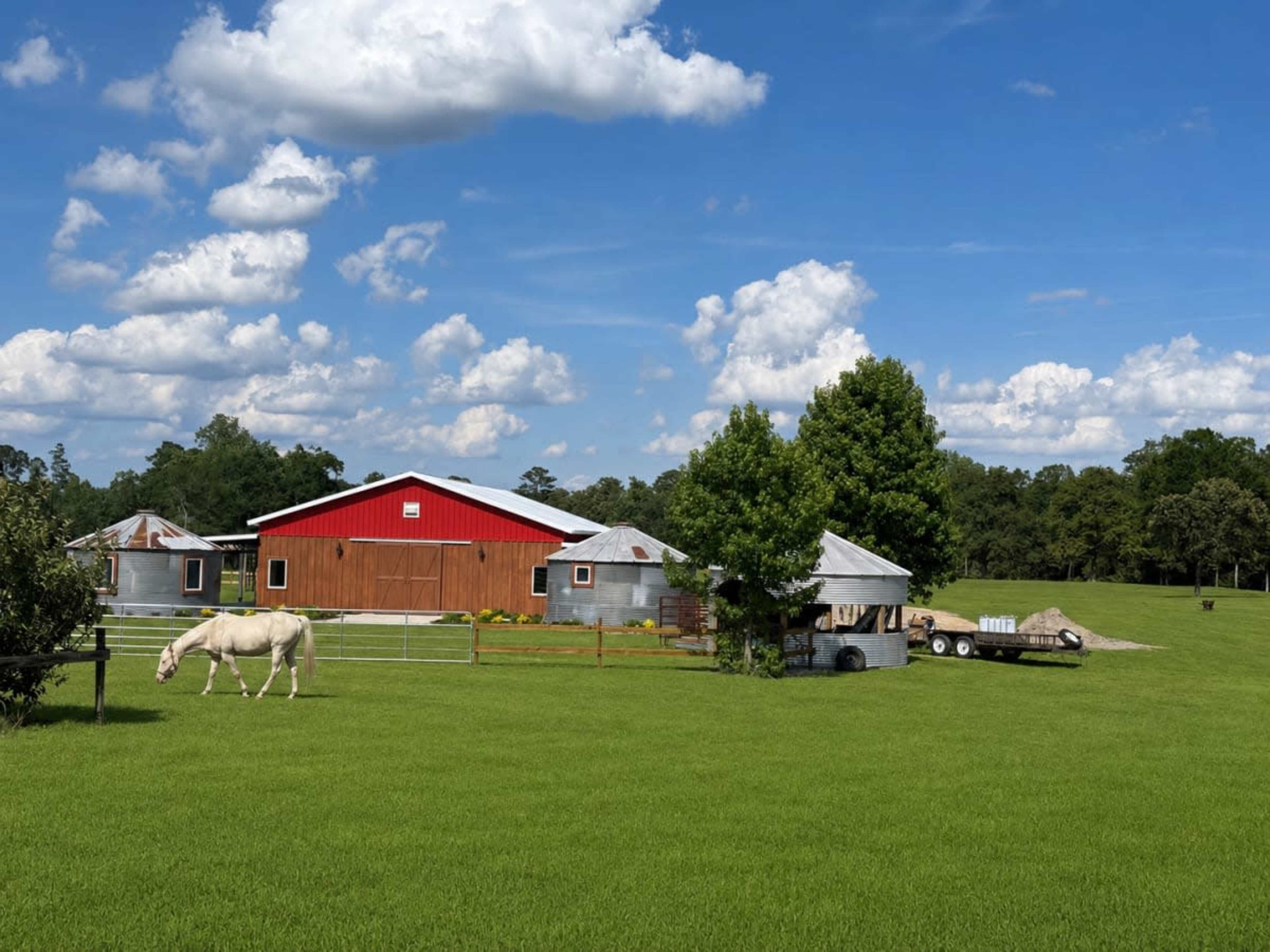 A red barn with corrugated metal buildings nearby is set against a blue sky with clouds, surrounded by green grass and a horse grazing in the field.