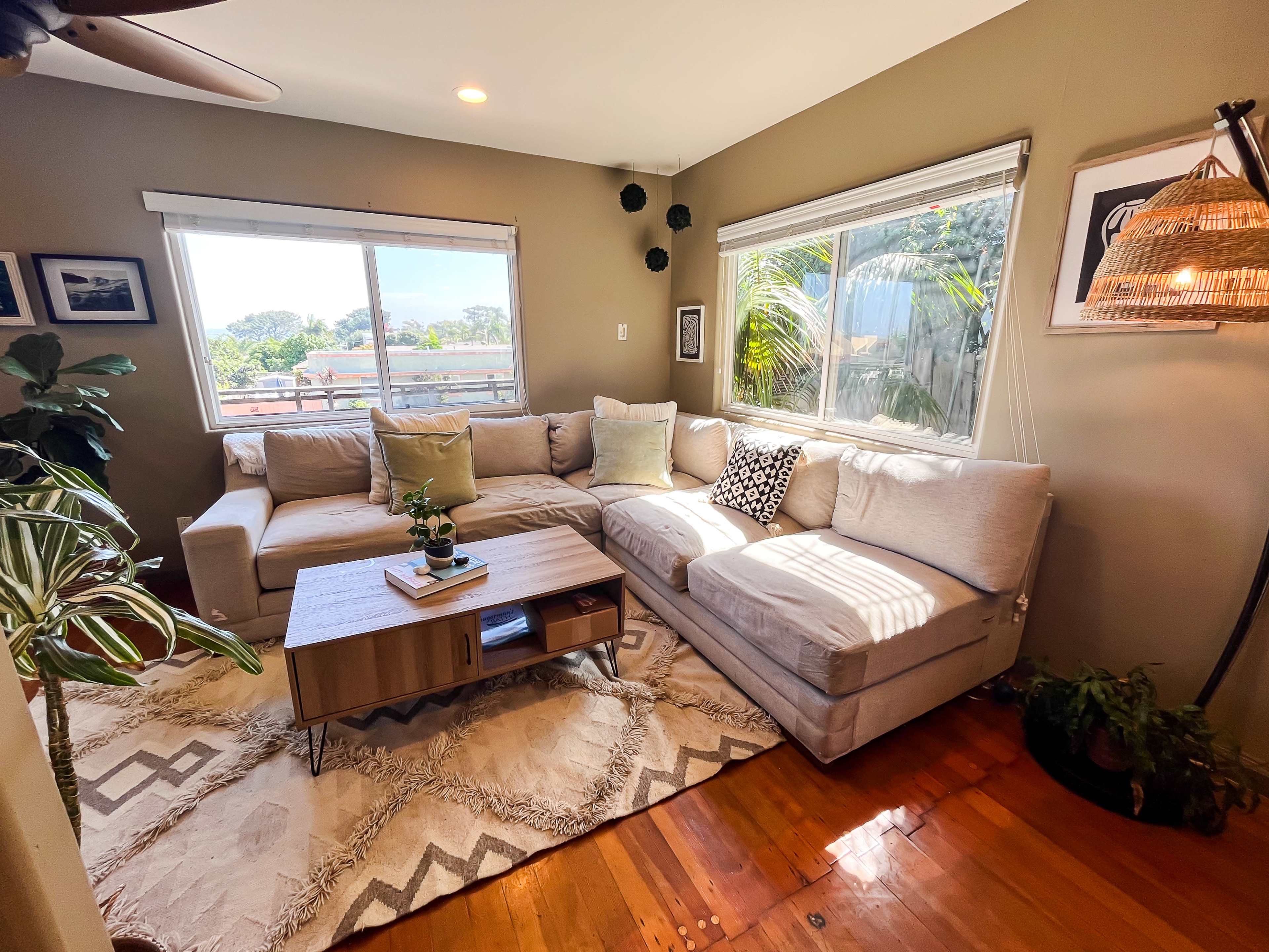 A light-colored sectional sofa positioned in a bright room with large windows, a wooden coffee table, and decorative plants.