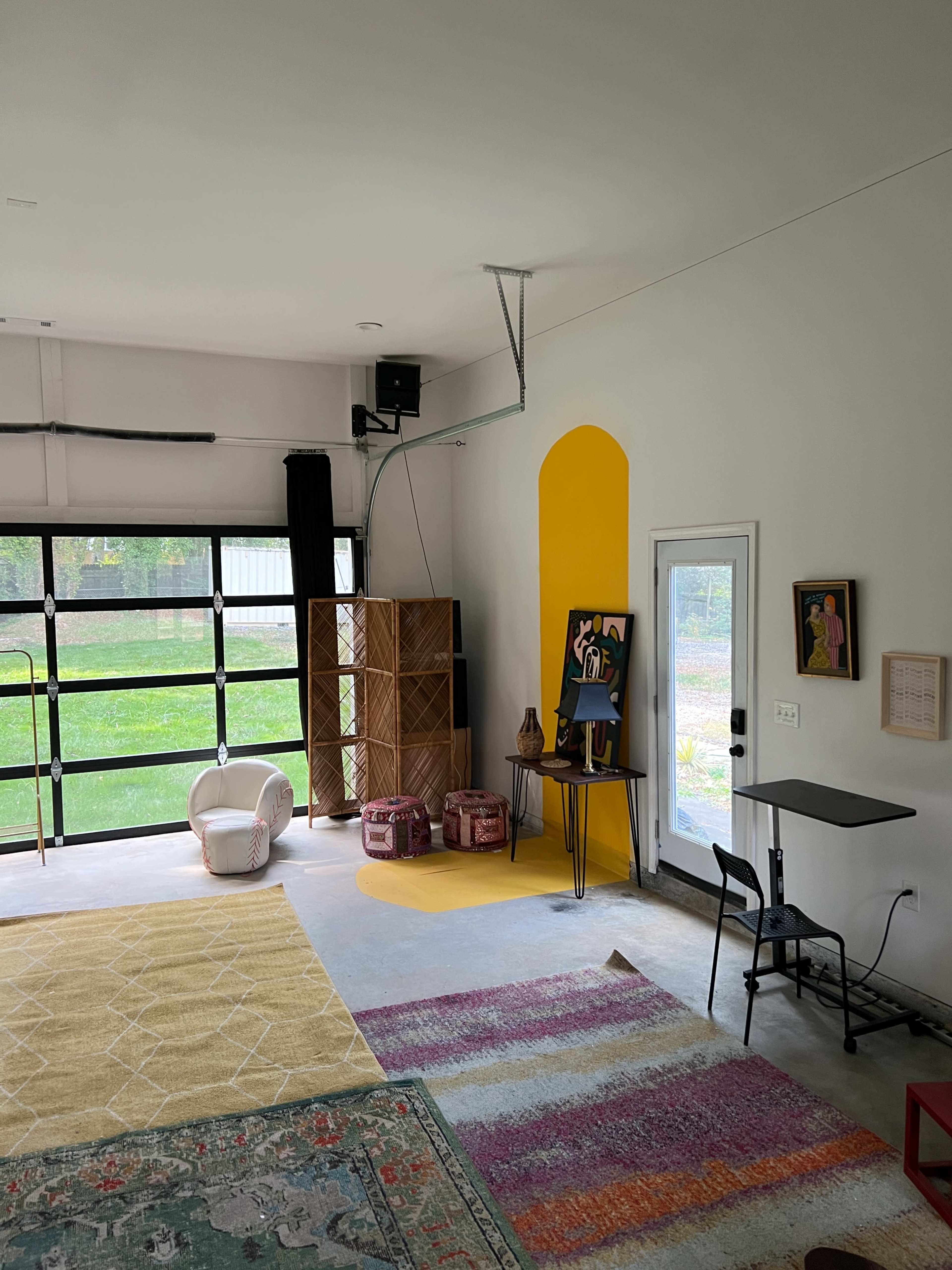 The image shows a spacious garage with a large glass garage door, a colorful area rug, furniture including a small table and a chair, and a bright yellow wall accent.