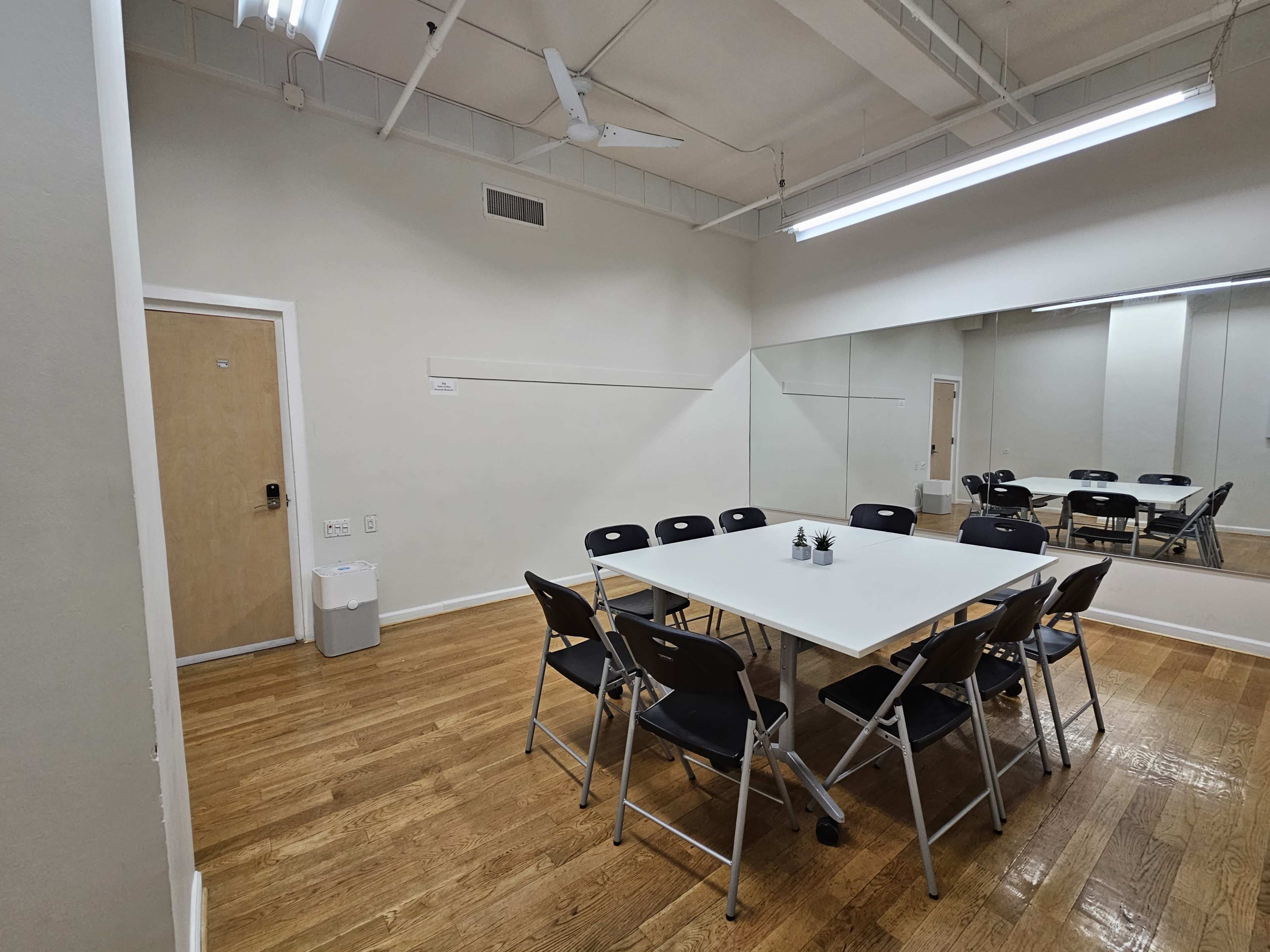 A meeting room with a large white table surrounded by black folding chairs, a door on one side, and a mirrored wall opposite the table.