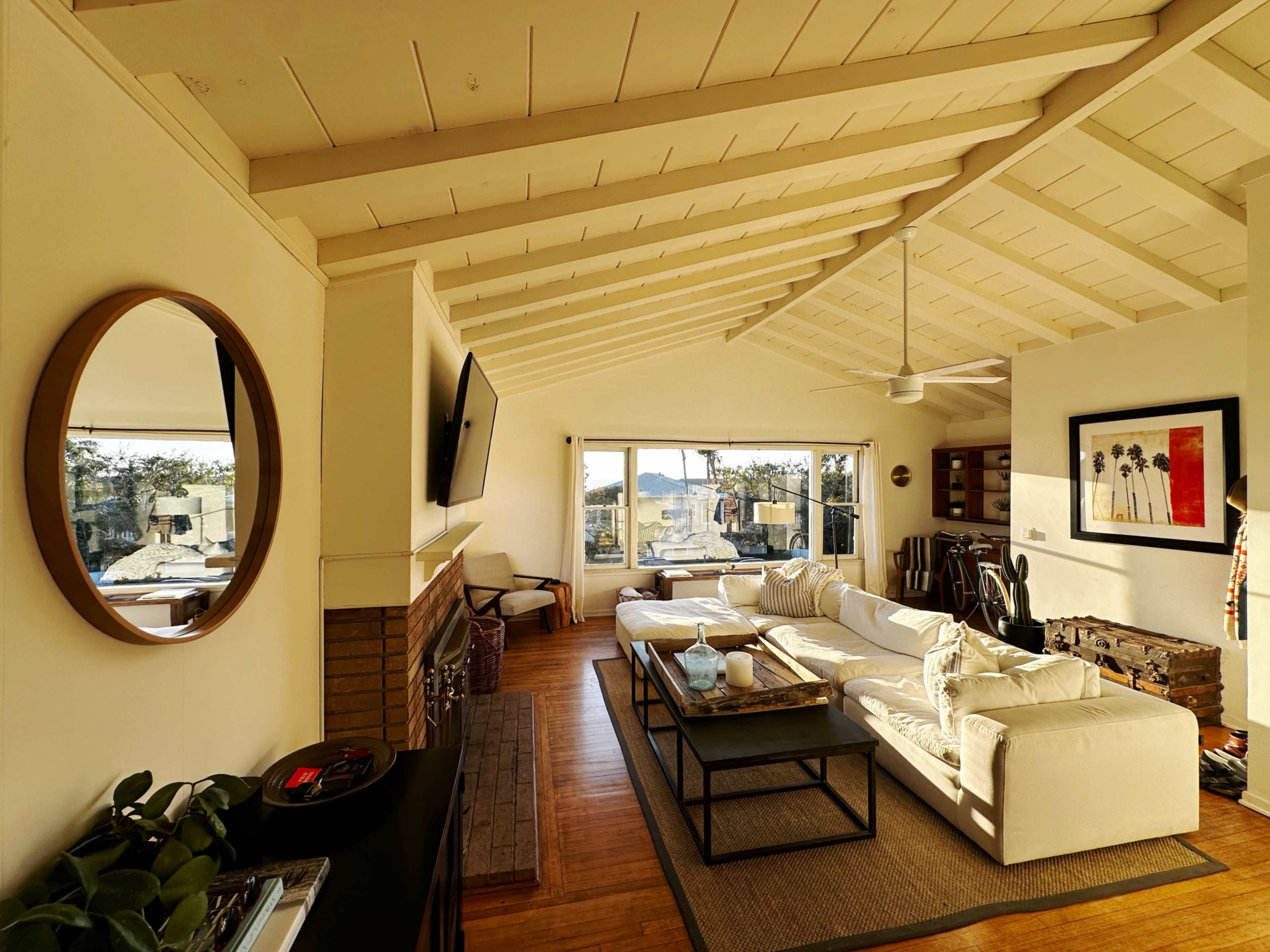 The image shows a spacious living room with high, beamed ceilings, a large window offering a view, a white sectional sofa, a coffee table, and a fireplace.