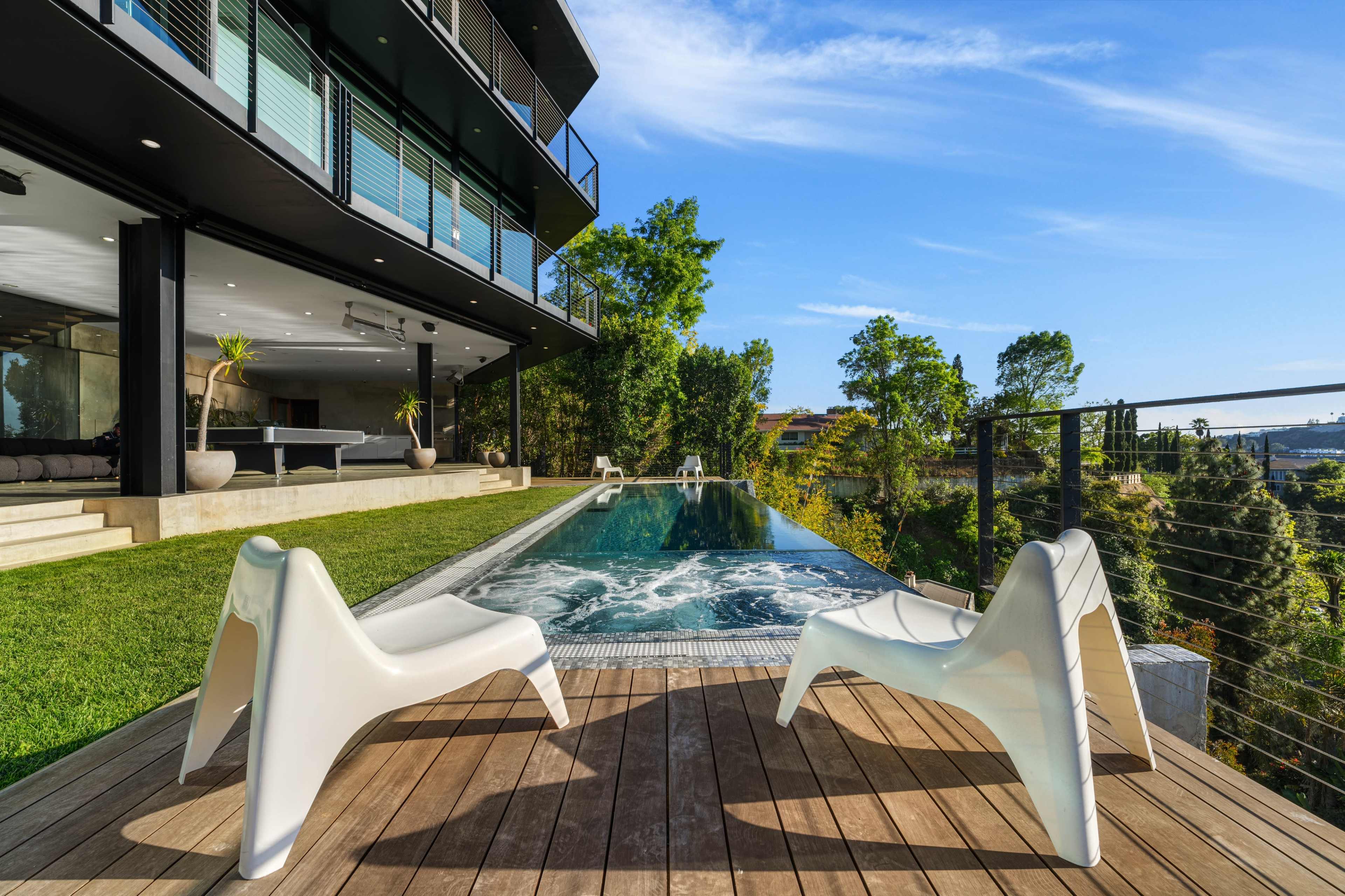 A modern building with large windows overlooks an infinity pool surrounded by wooden decking and lounge chairs.