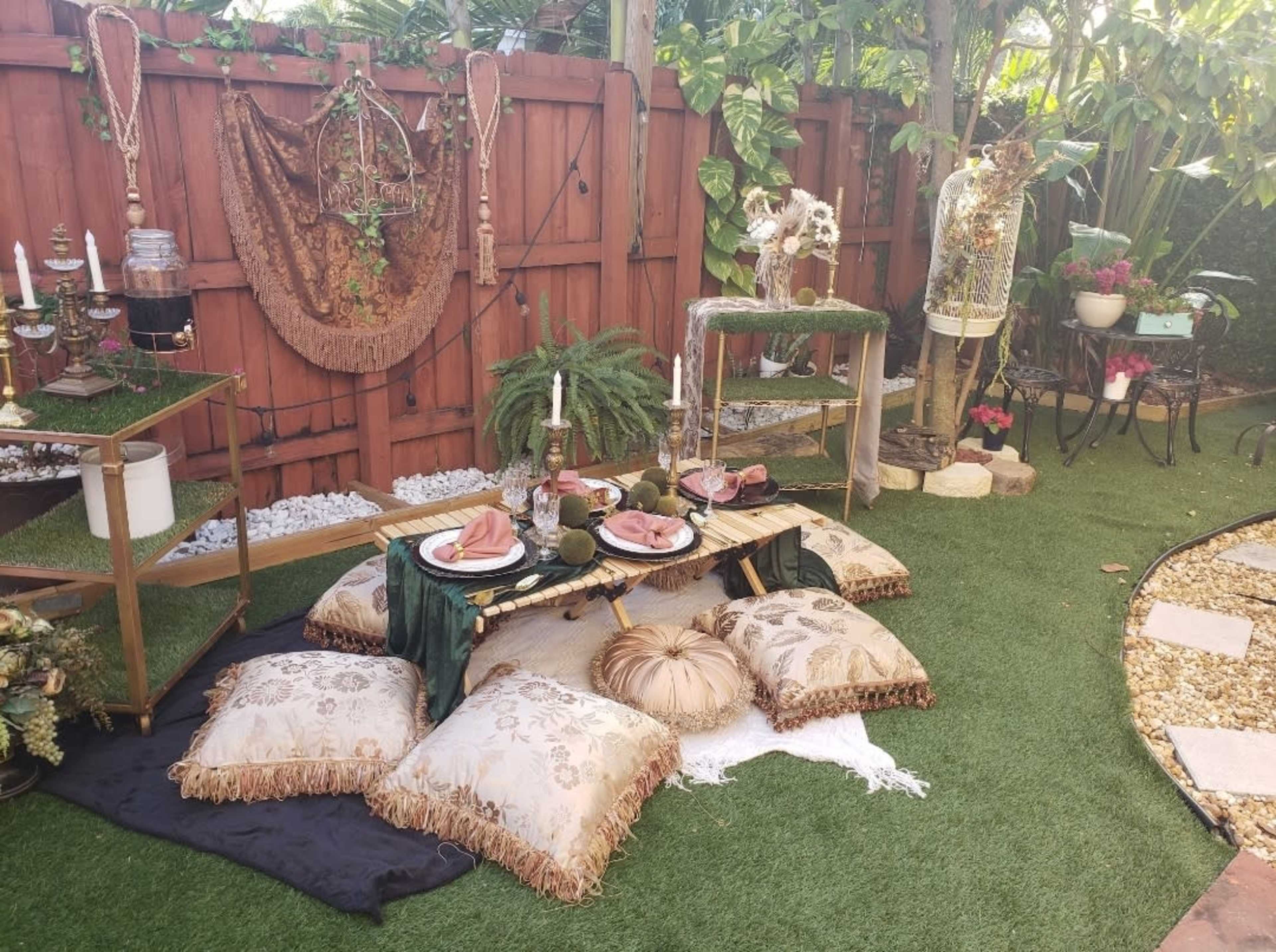 The scene features an outdoor setting with a picnic area covered in decorative pillows, a low table set with plates and food, surrounded by greenery and adorned with various decorative items.