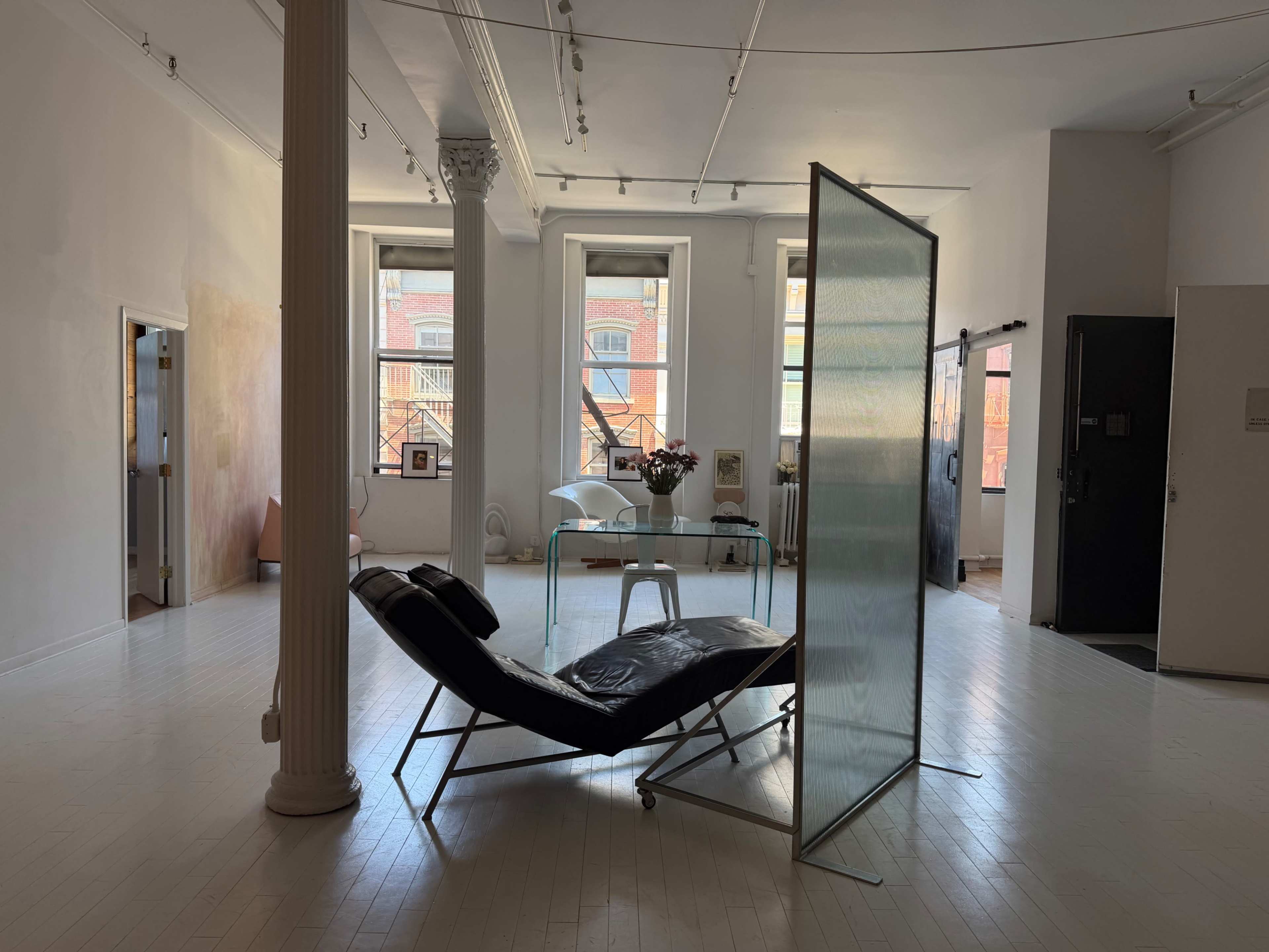 The image shows a spacious, minimalist interior with large windows, a black reclining chair, a glass partition, and a small table with a floral arrangement.