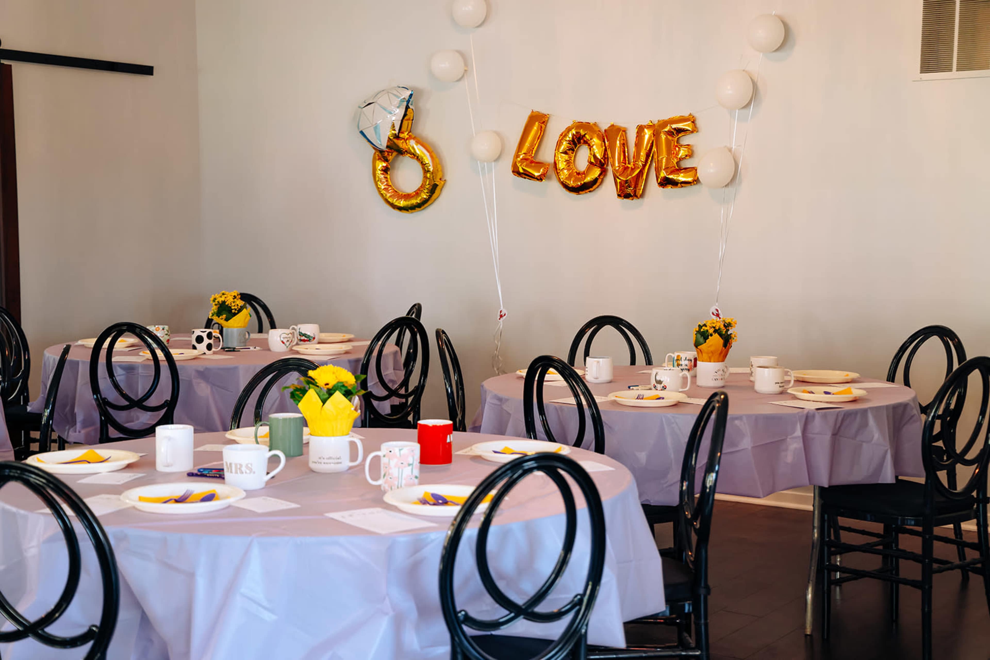 A decorated room features several round tables set with tablecloths, mugs, and floral centerpieces, with a balloon display spelling "LOVE" on the wall.