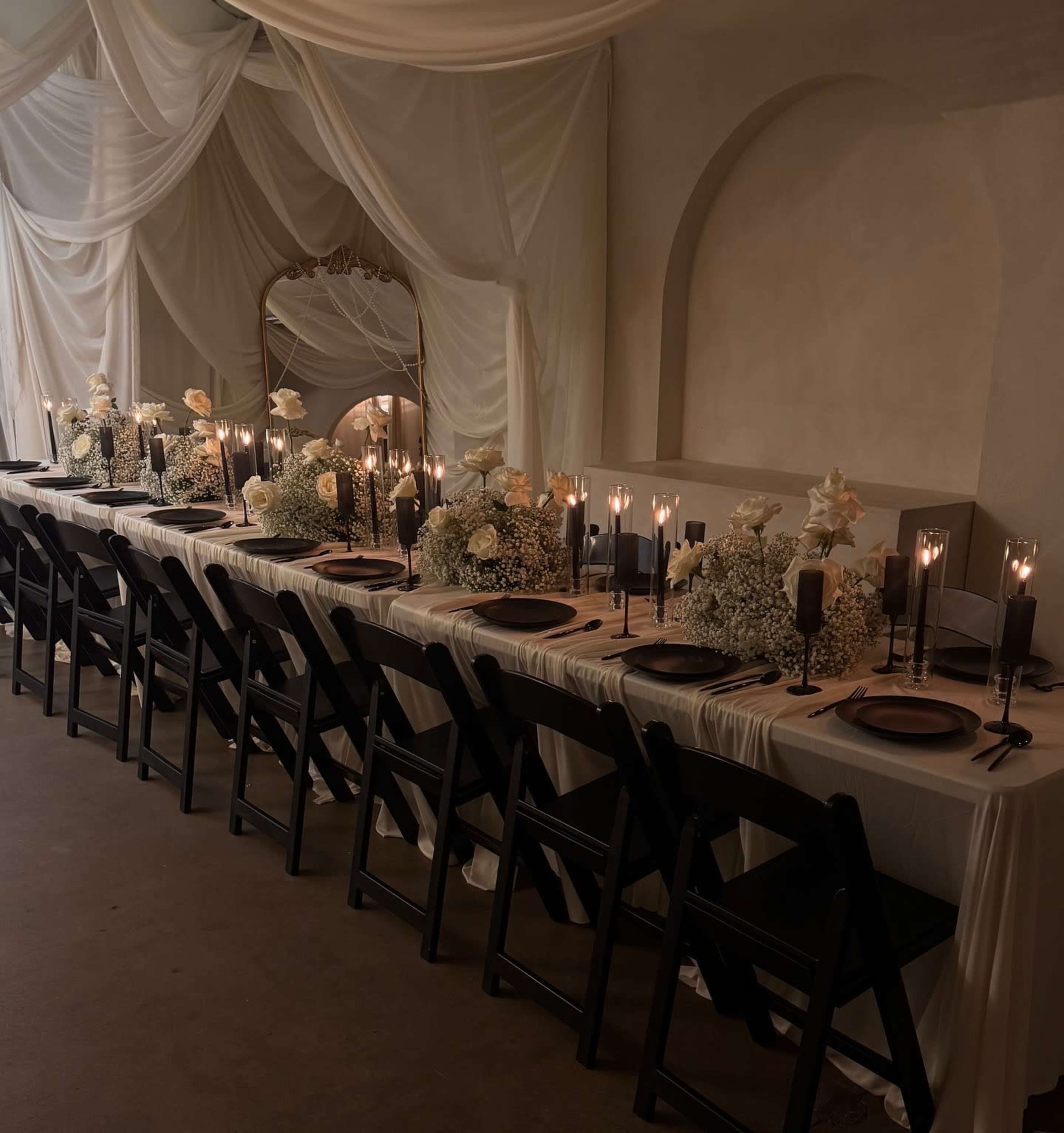 A long dining table is elegantly set with dark plates, white flowers, and candlelight in a softly draped room.