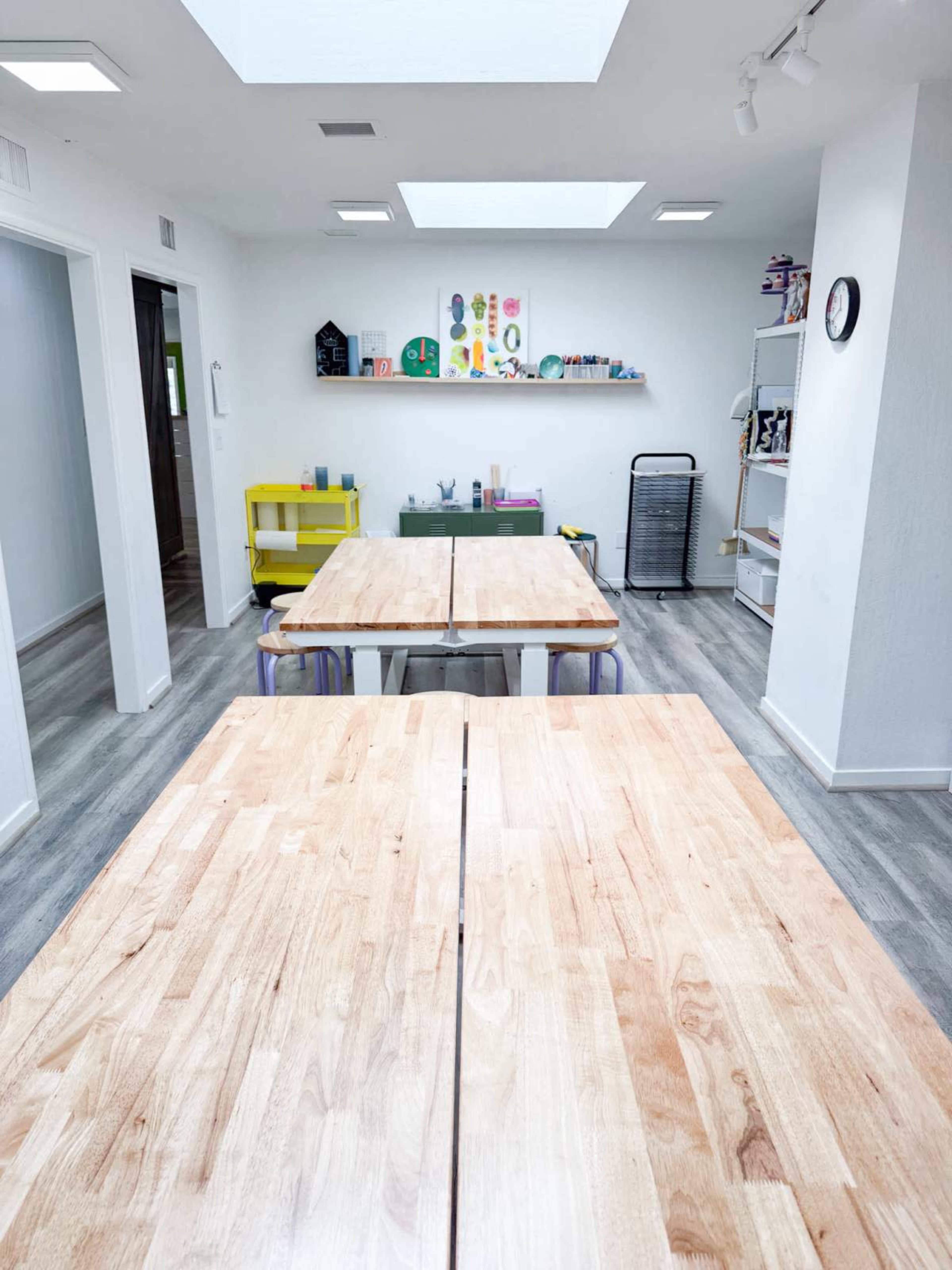 A bright, airy room with two wooden tables, a yellow shelf, and various supplies neatly organized on shelves against the walls.