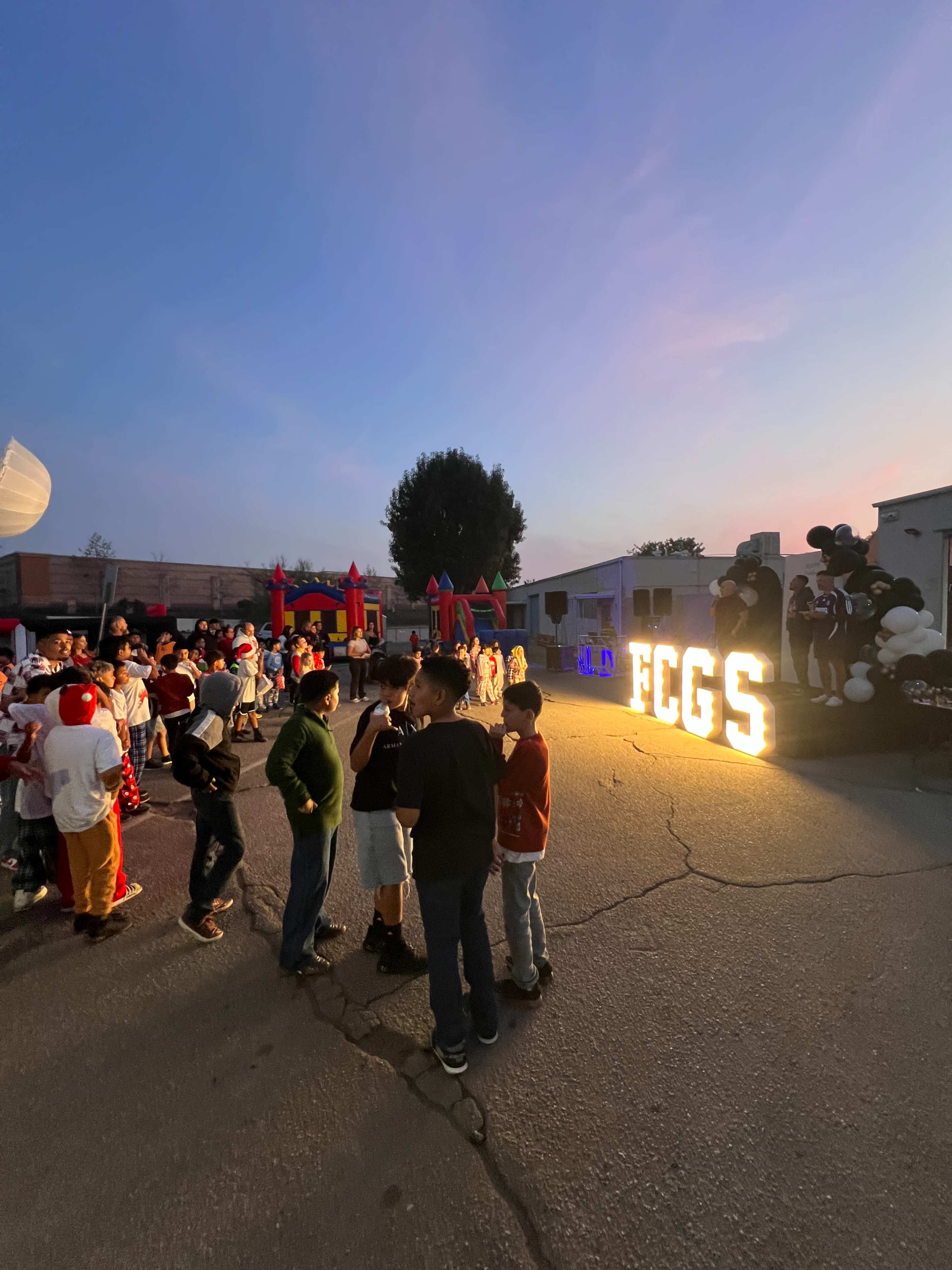 A gathering of people in costumes stands near illuminated letters spelling "FCGS" at an outdoor event with inflatable attractions in the background during twilight.