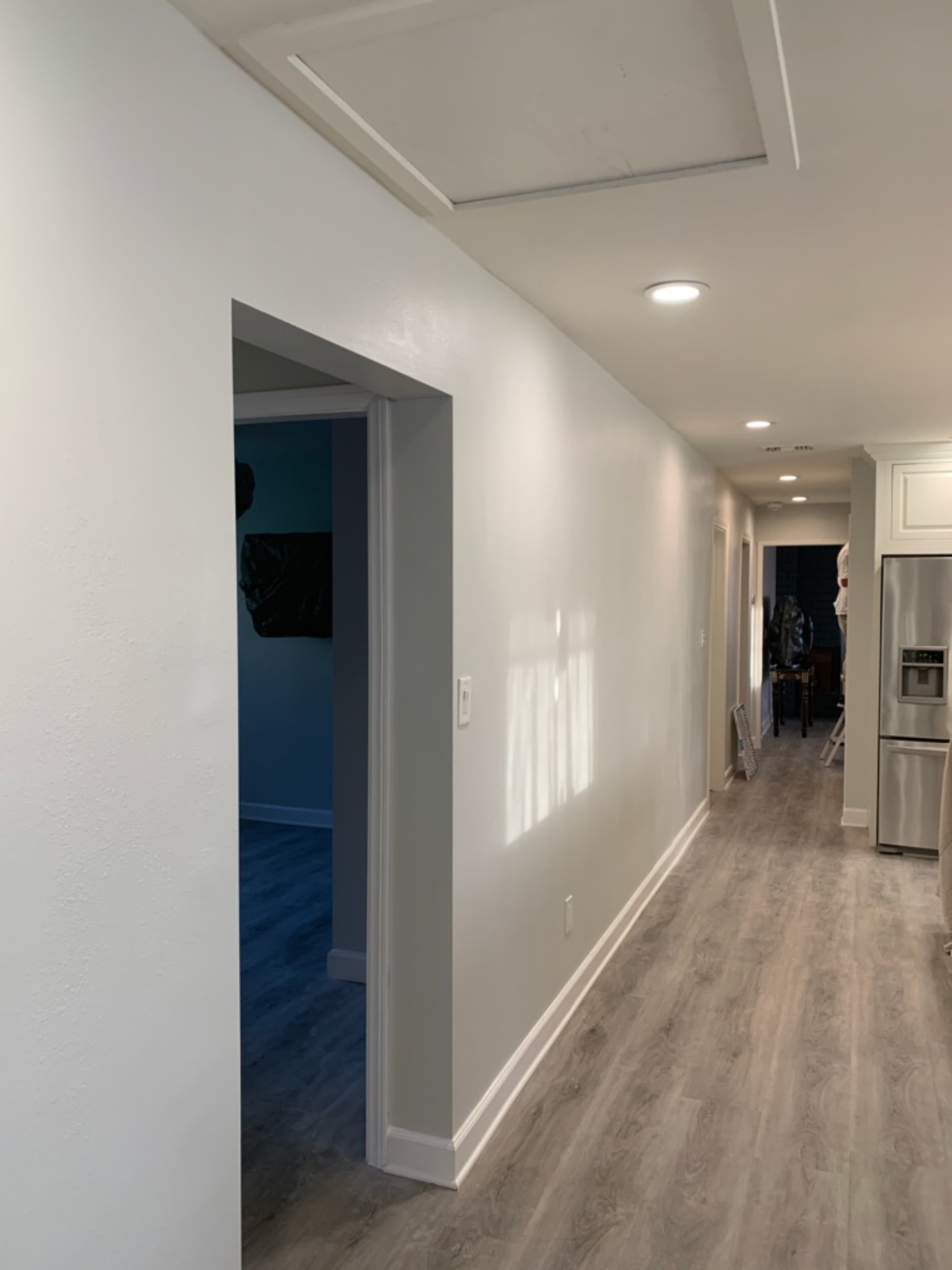 A hallway with light-colored walls and laminate flooring runs alongside an open doorway leading to another room.