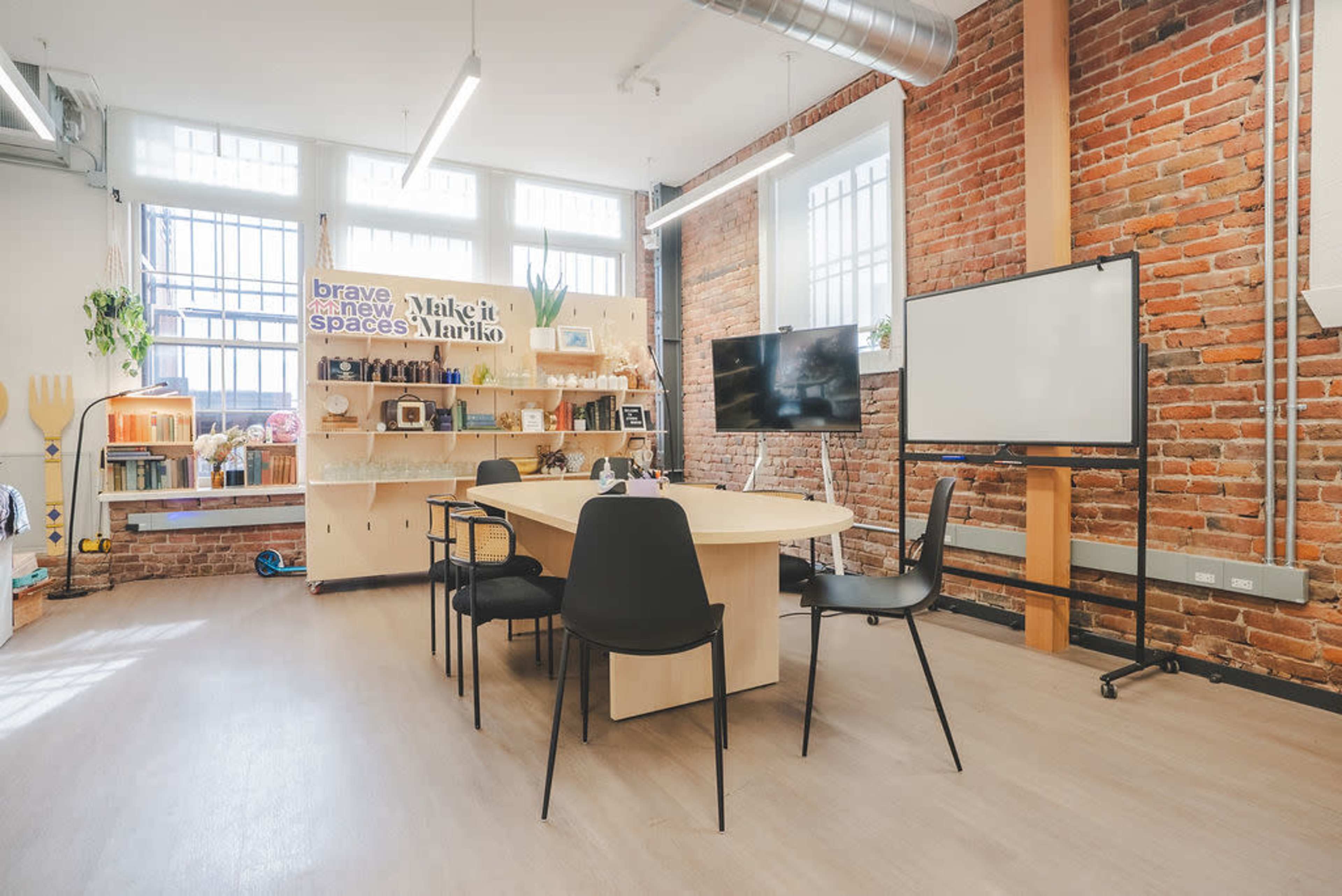 The image shows a modern meeting room with a large table, several black chairs, exposed brick walls, and a whiteboard, along with shelves displaying various items and decor.