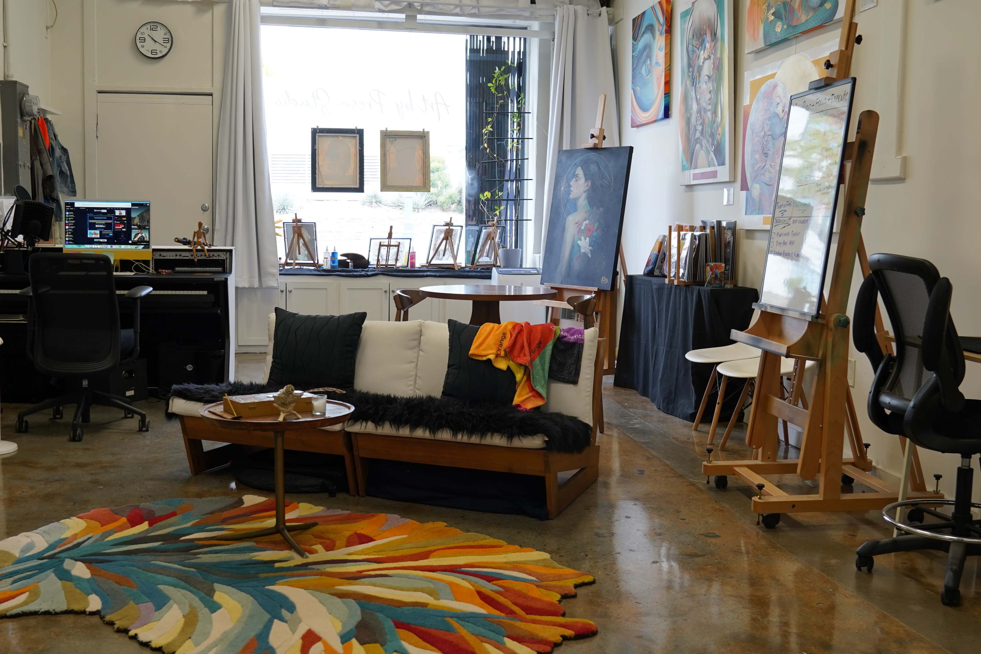 The image shows an artist's studio featuring a colorful area rug, a wooden coffee table with decor, a black couch with cushions, and various artworks displayed on the walls.