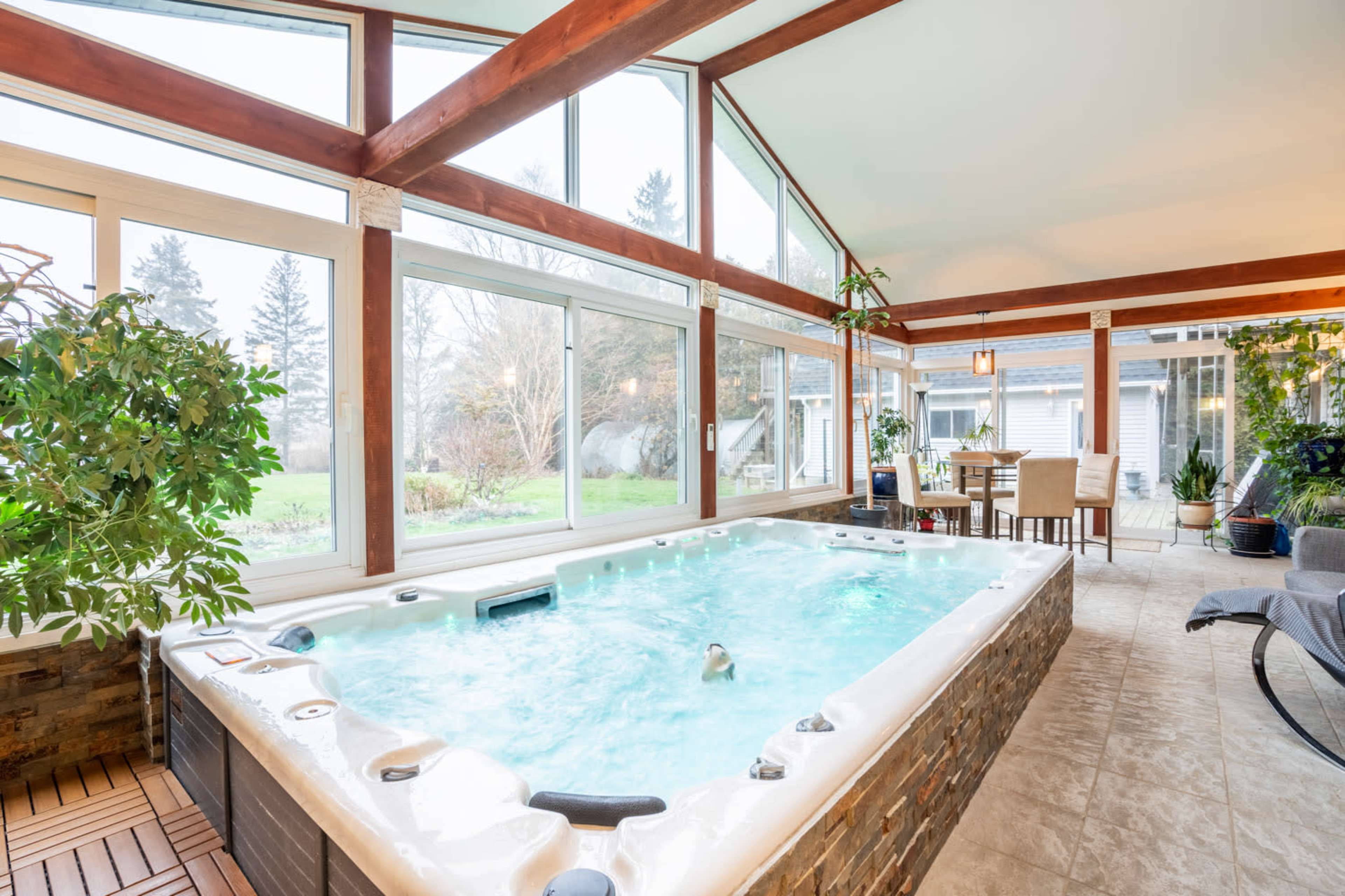 The image shows an indoor room featuring a hot tub surrounded by large windows, potted plants, and a dining area.