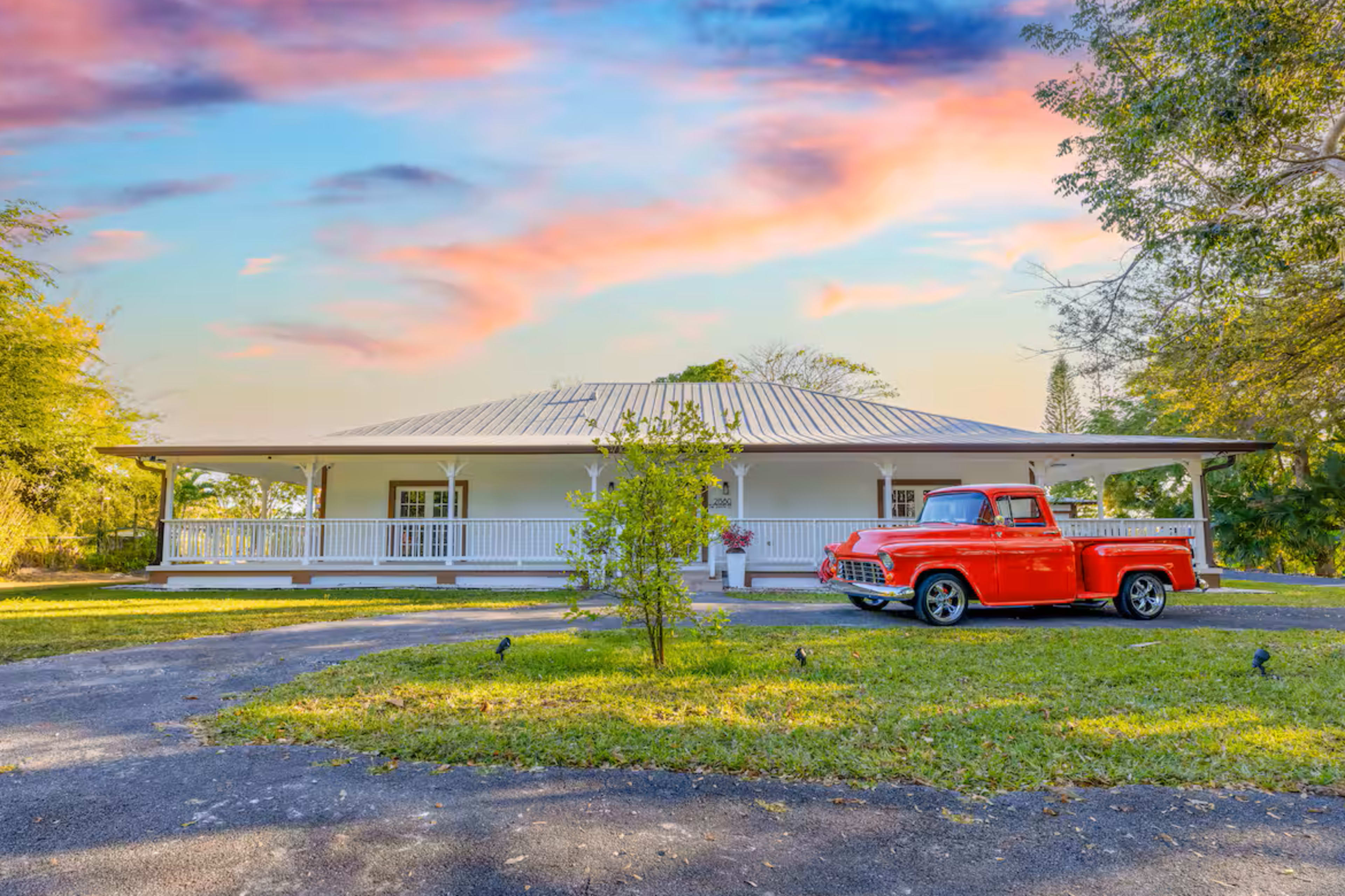 A vintage red truck is parked in front of a white house with a wraparound porch at sunset.