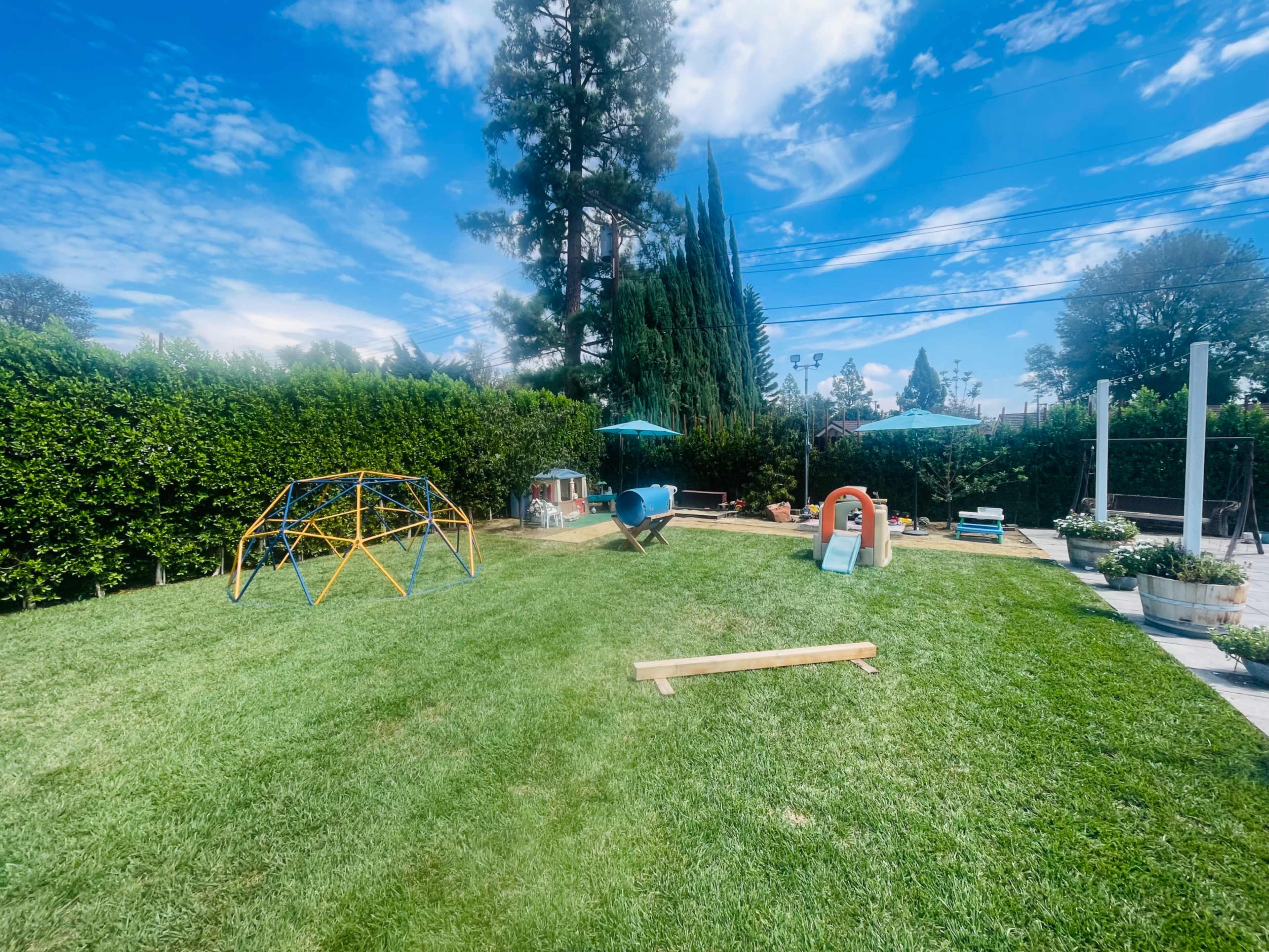 A backyard play area featuring a colorful climbing structure, a slide, and seating under green umbrellas, all set on a neatly maintained lawn.