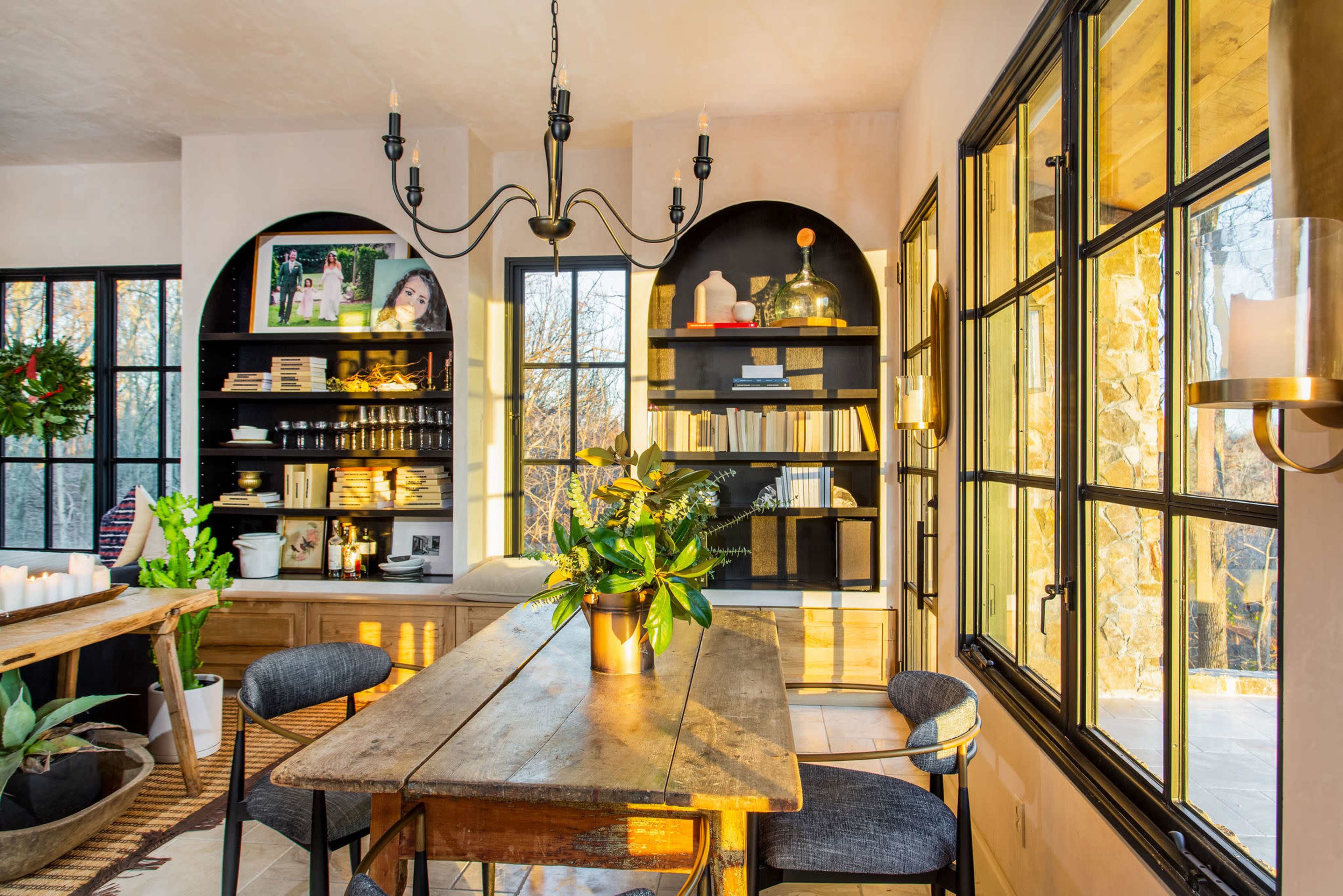 A well-lit dining area features a wooden table surrounded by chairs, with shelves displaying books and decorative items against a backdrop of large windows.