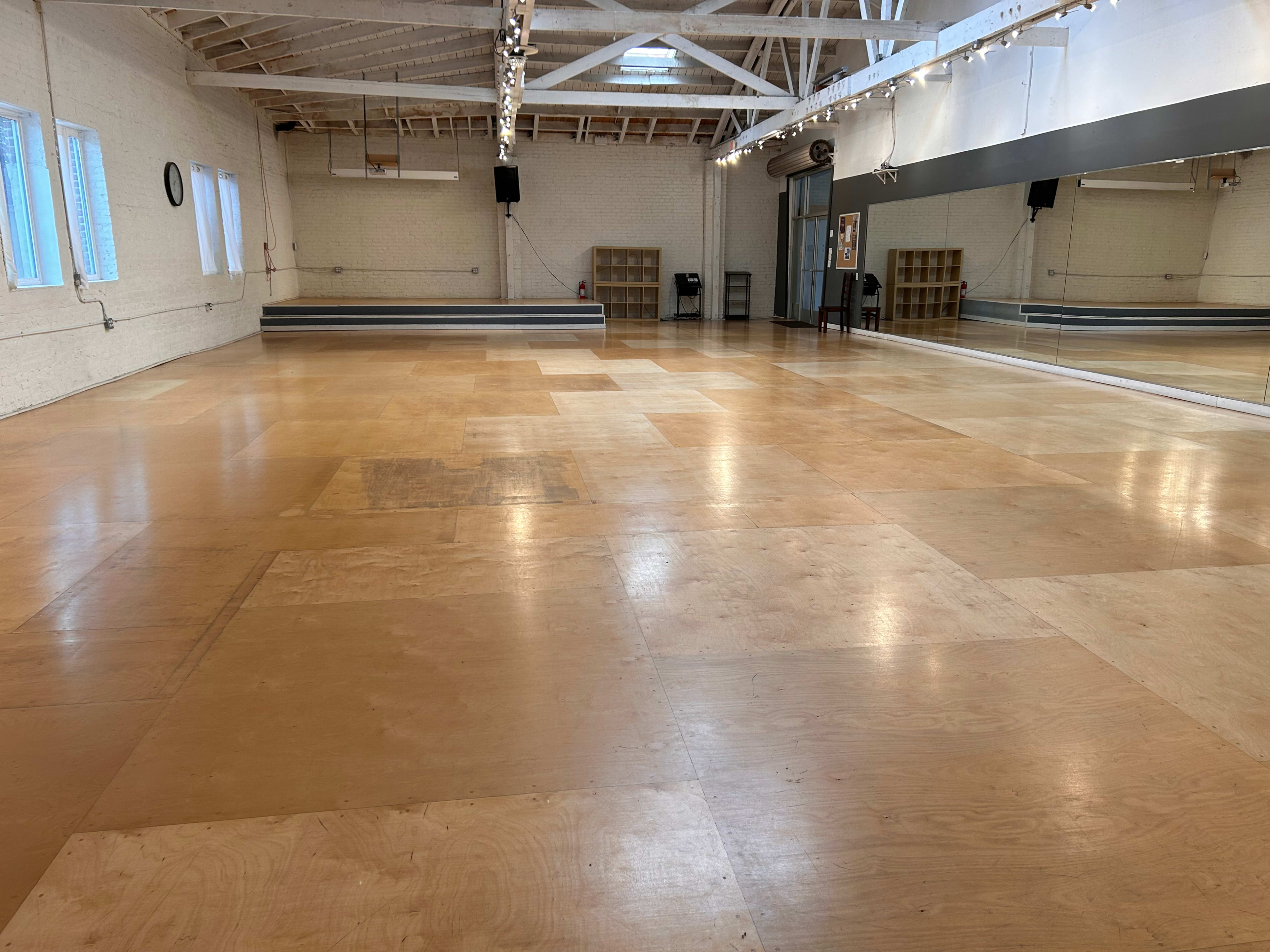 A spacious dance studio with wooden flooring, large mirrors along one wall, and natural light coming through several windows.