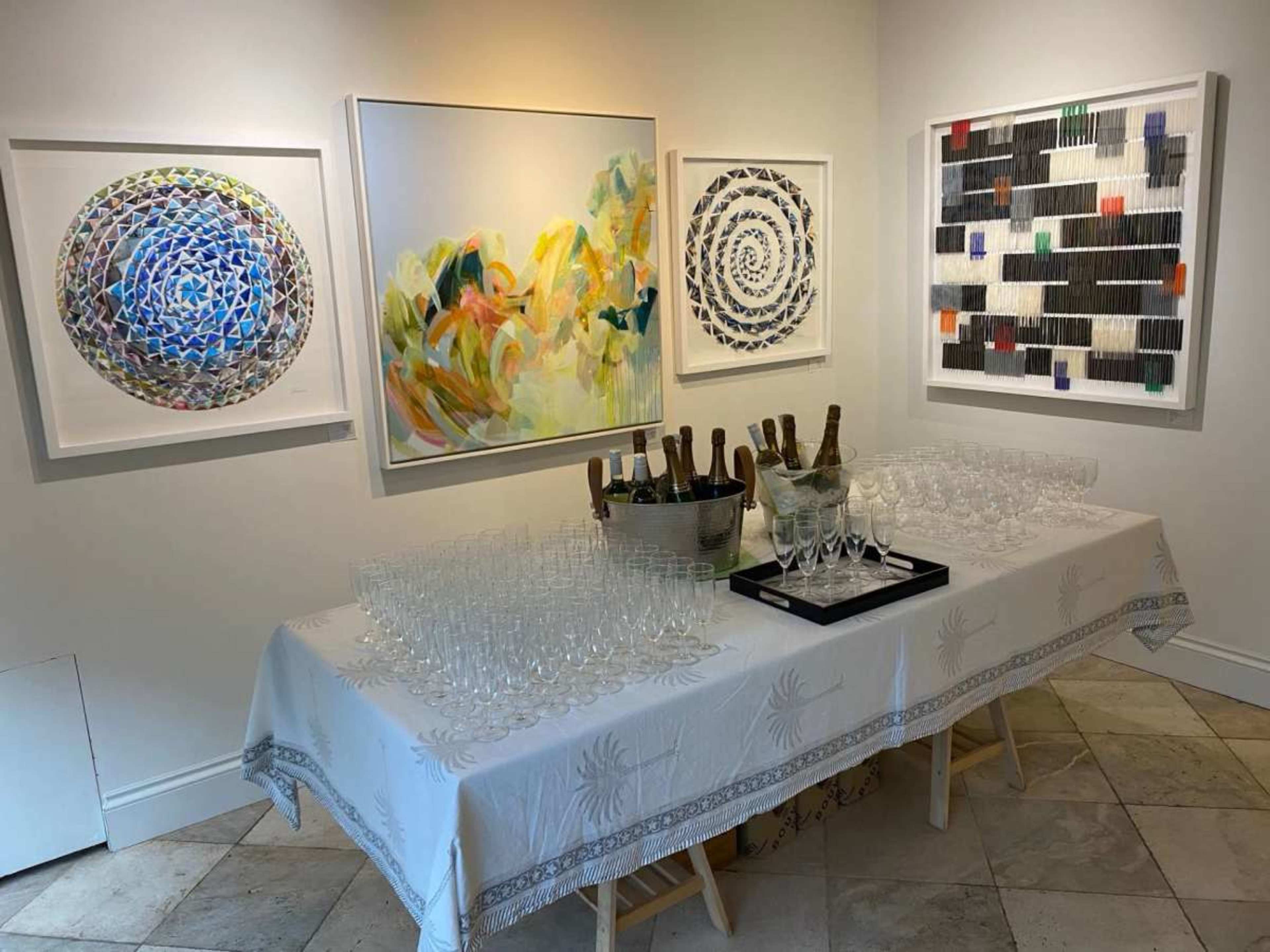 The image shows a table covered with a white tablecloth holding numerous empty glasses and bottles, set against a wall adorned with four colorful art pieces.