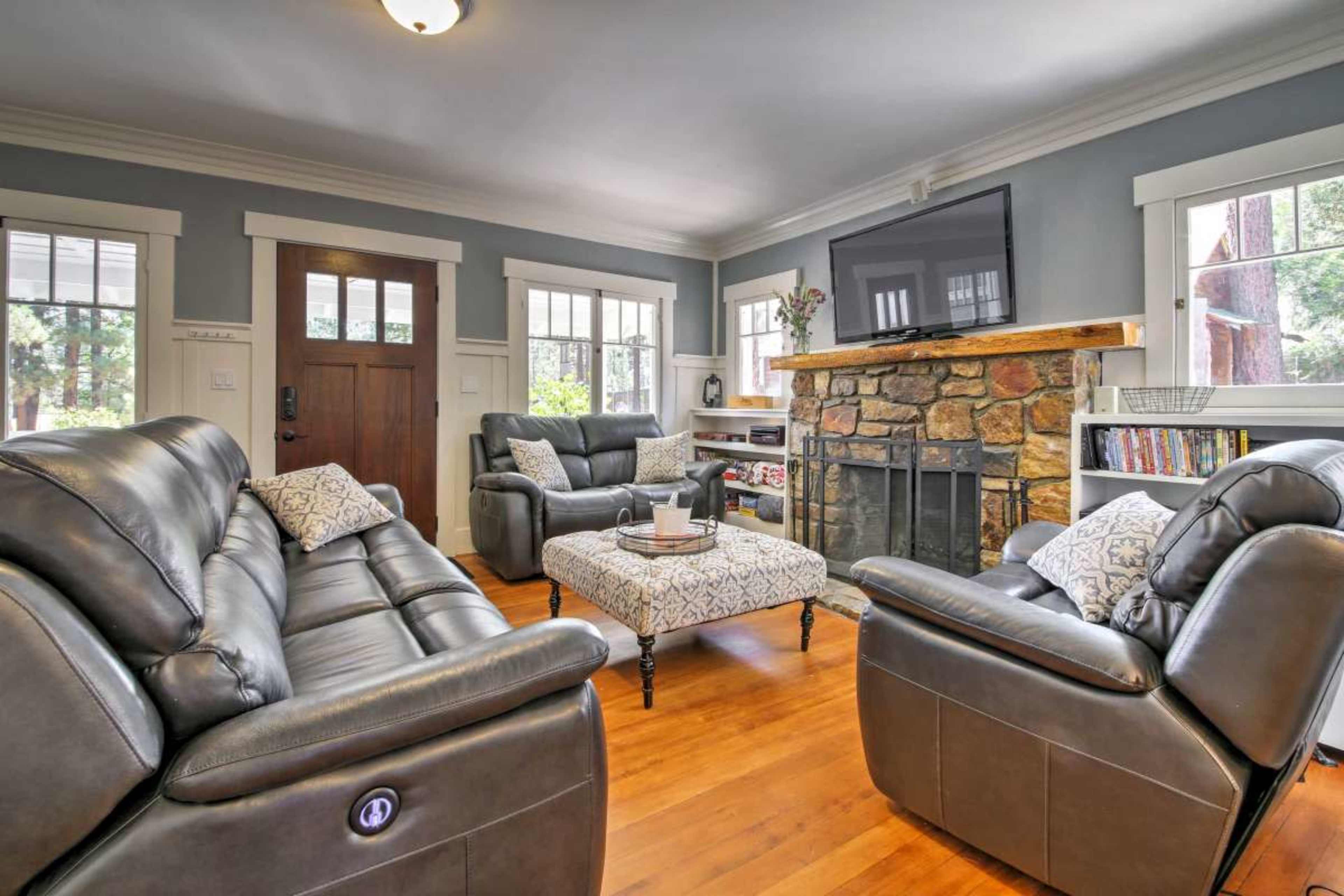 A living room featuring leather chairs and a couch, a stone fireplace, and a large television mounted on the wall, with wood flooring and natural light from several windows.