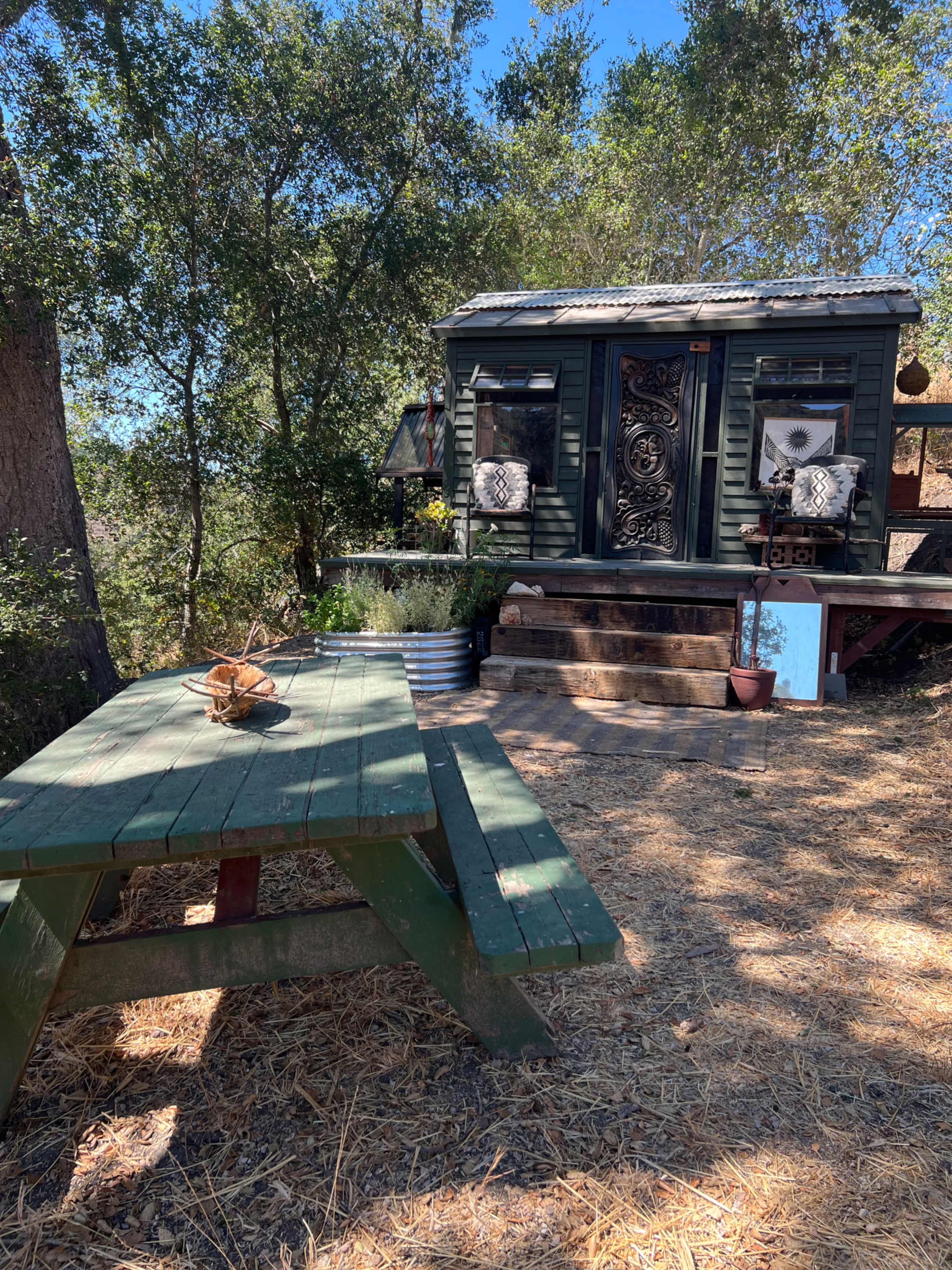 The image shows a rustic green picnic table in front of a small green cabin surrounded by trees and natural landscaping.