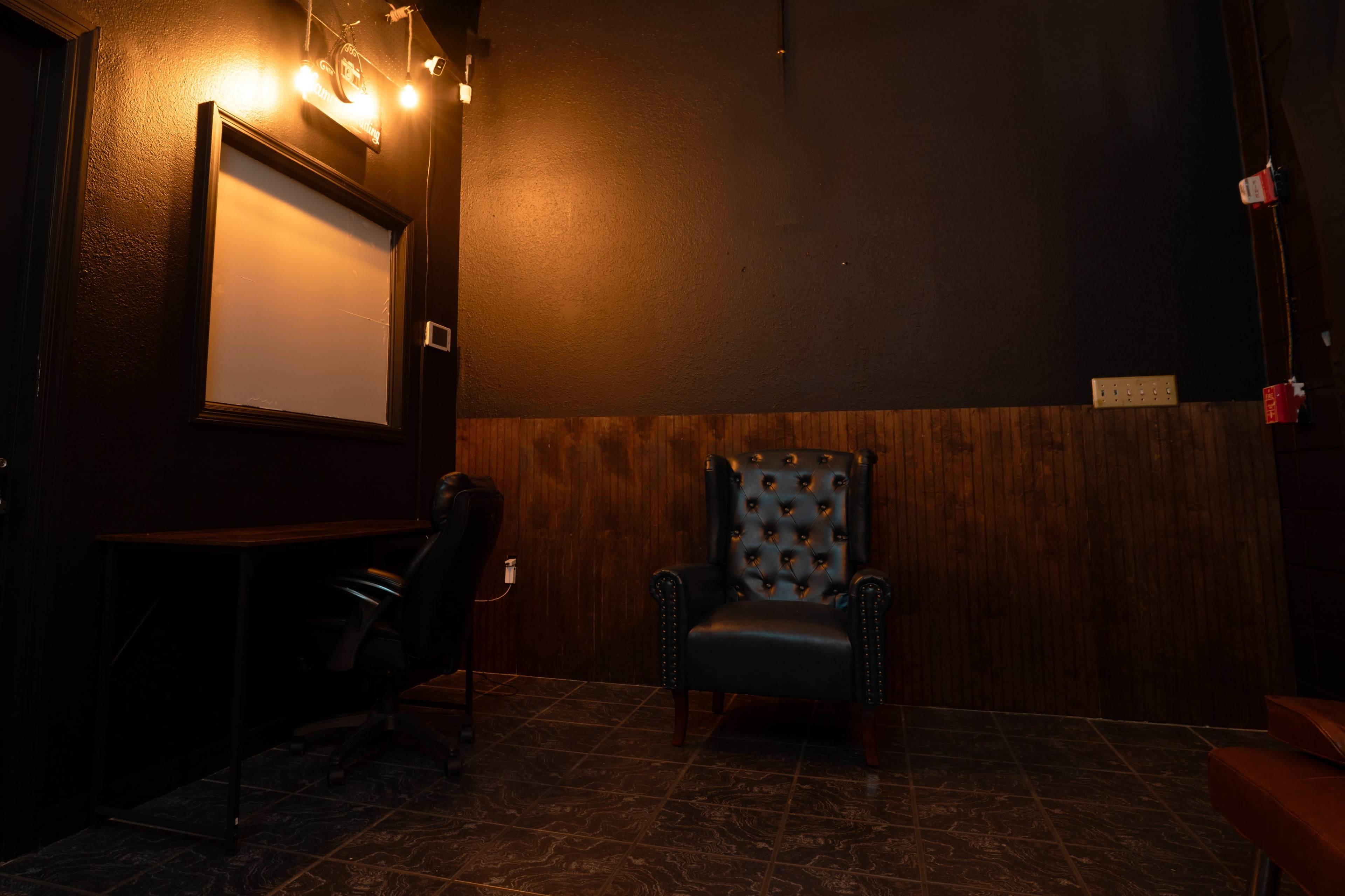 The image shows a dimly lit room featuring a black leather chair, a wooden wall panel, a desk with a swivel chair, and wall-mounted lights.
