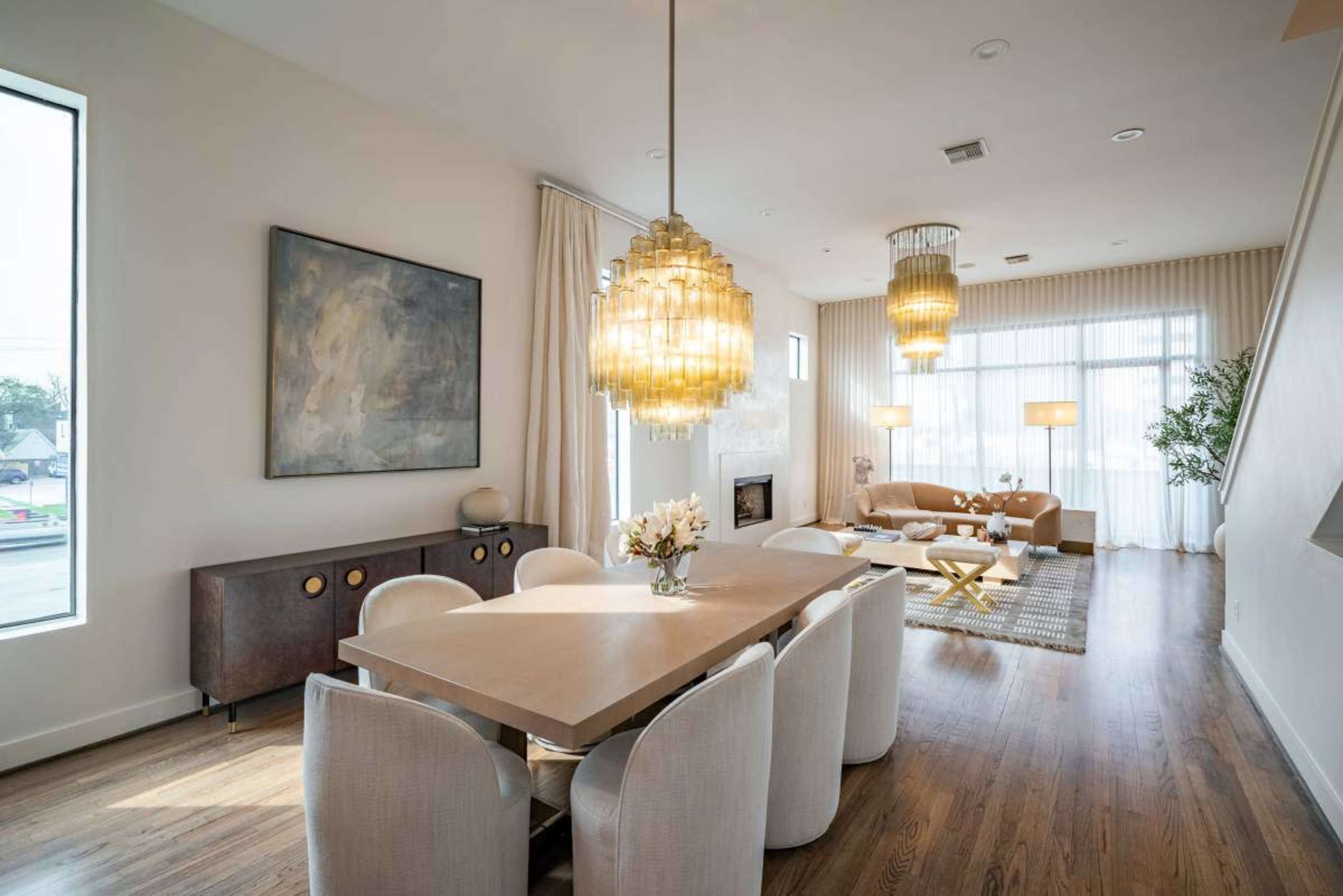 The image showcases a modern dining room featuring a light wood table surrounded by upholstered chairs, with large pendant lights and a cozy seating area in the background.