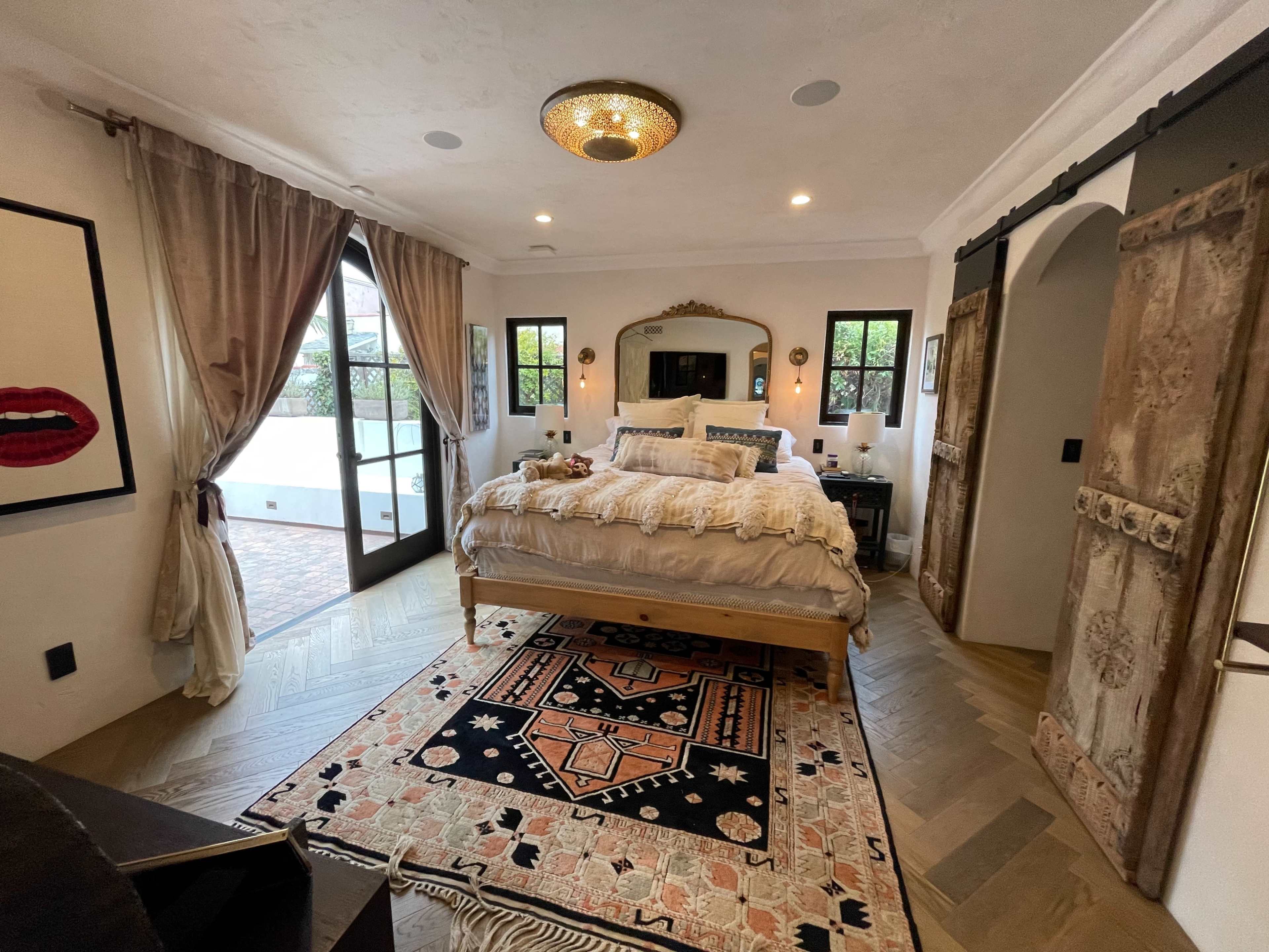 A well-decorated bedroom featuring a large bed, a decorative rug, a mirror, and double doors leading to an outdoor area.