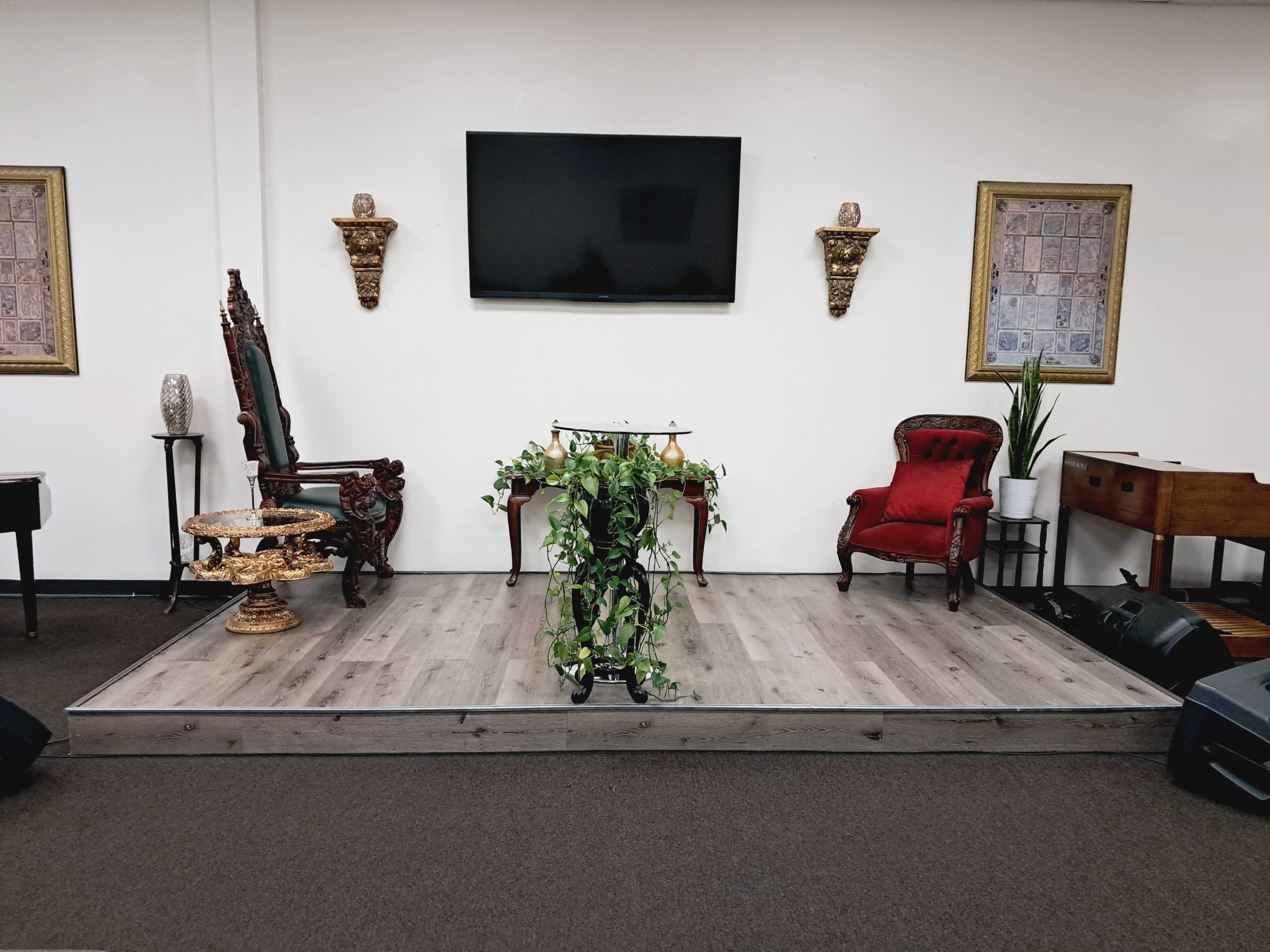 The image shows a stage with two ornate chairs, a decorative table, a green plant, and a television mounted on the wall, all within a room featuring a simple, neutral-colored background.