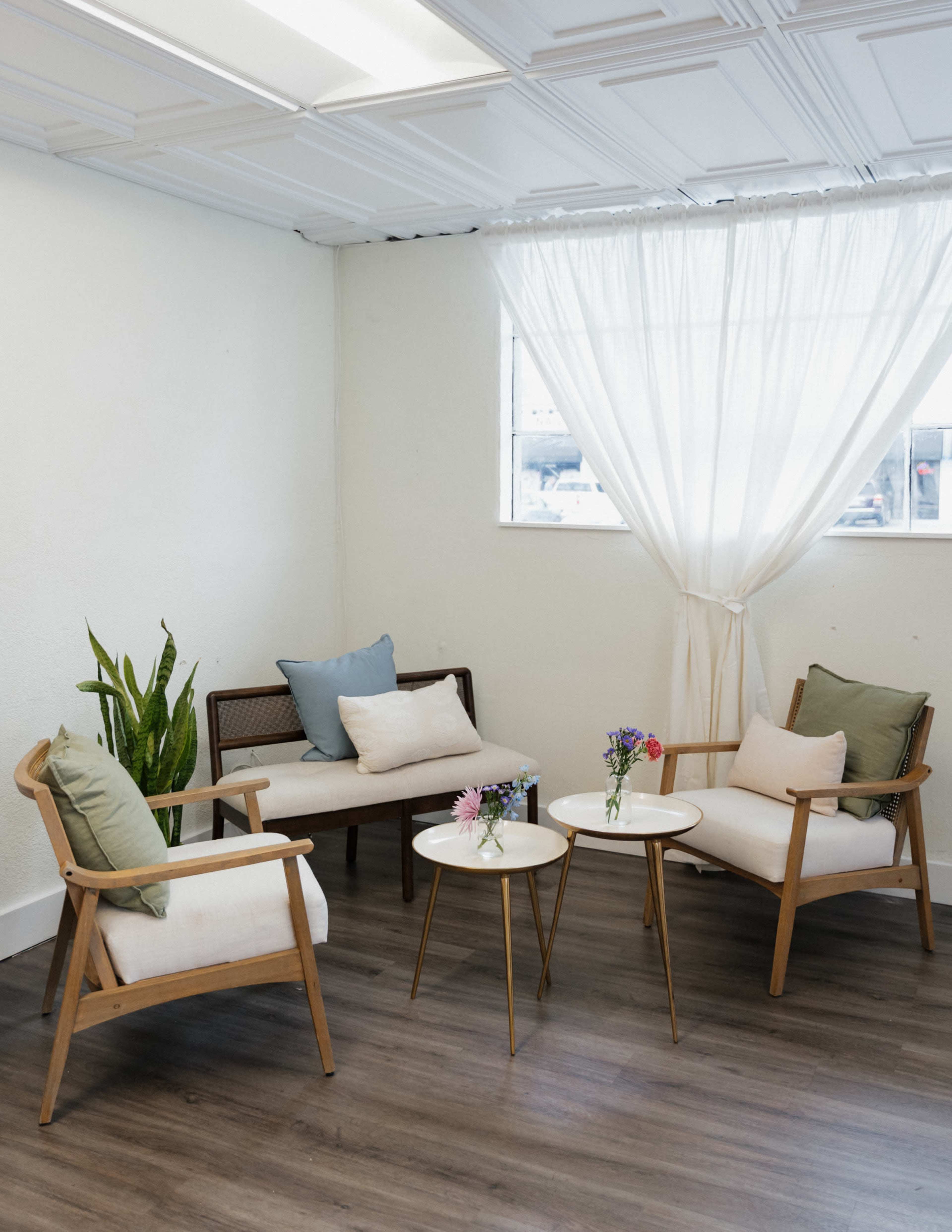 The image shows a small, minimalist seating area with two wooden armchairs and a sofa, accompanied by two circular coffee tables and a potted plant, all situated near a window with sheer curtains.