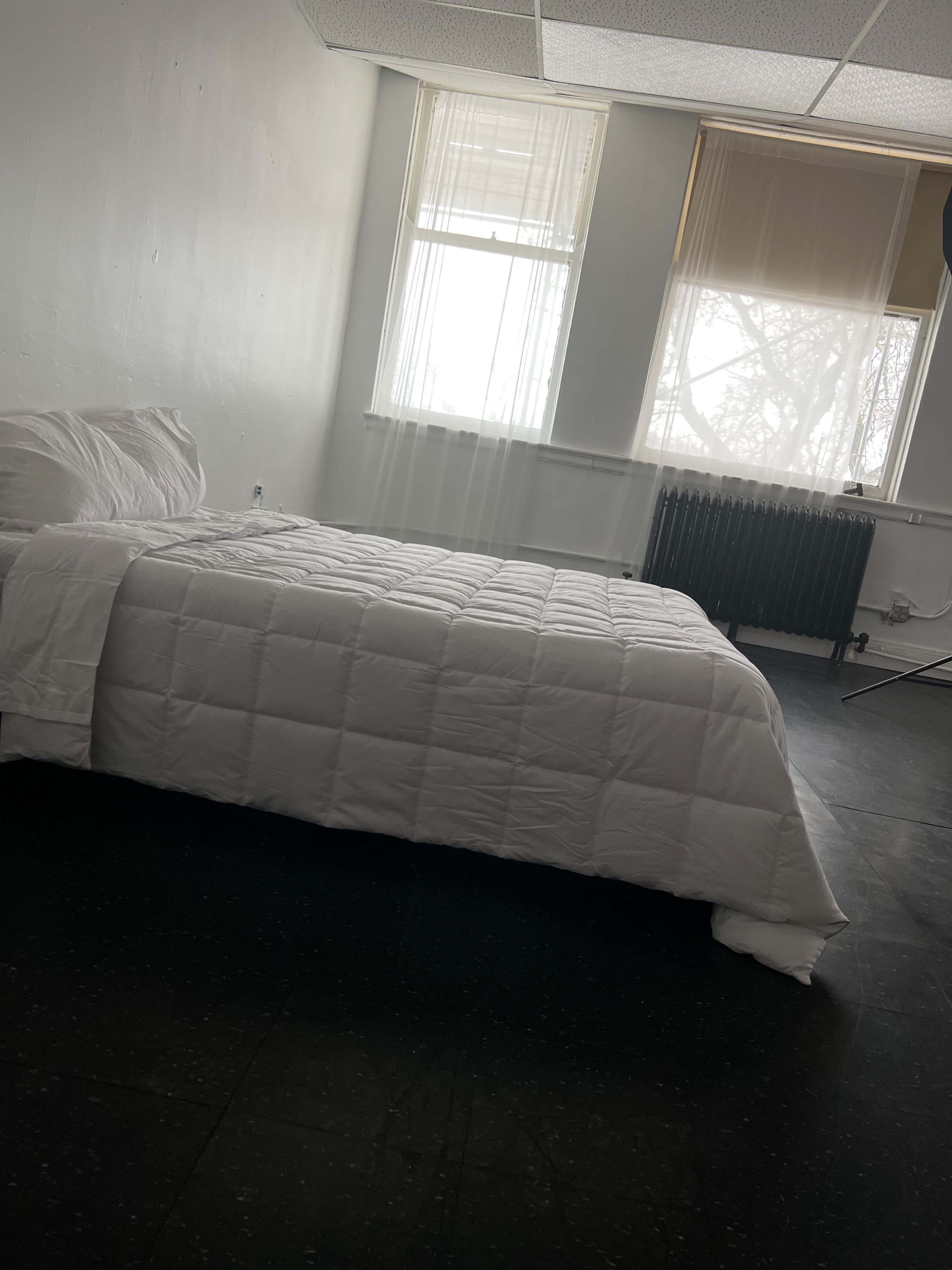 A minimalistic room with a bed covered in a white quilt, two windows letting in light, and a radiator against the wall.