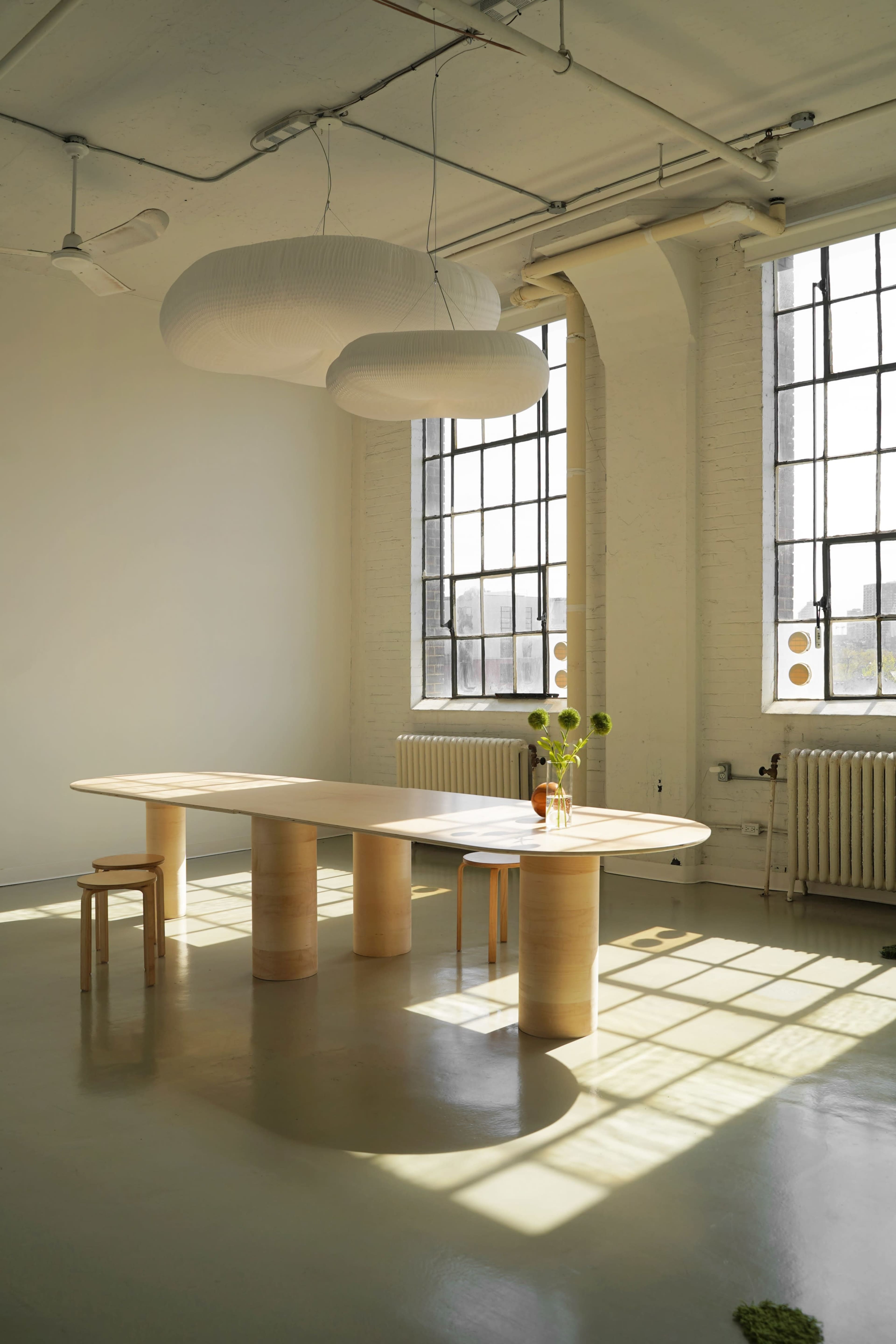 A modern, minimalist dining area features a long wooden table with cylindrical legs, paired with two simple wooden chairs, under soft hanging lights, illuminated by sunlight streaming through large windows.