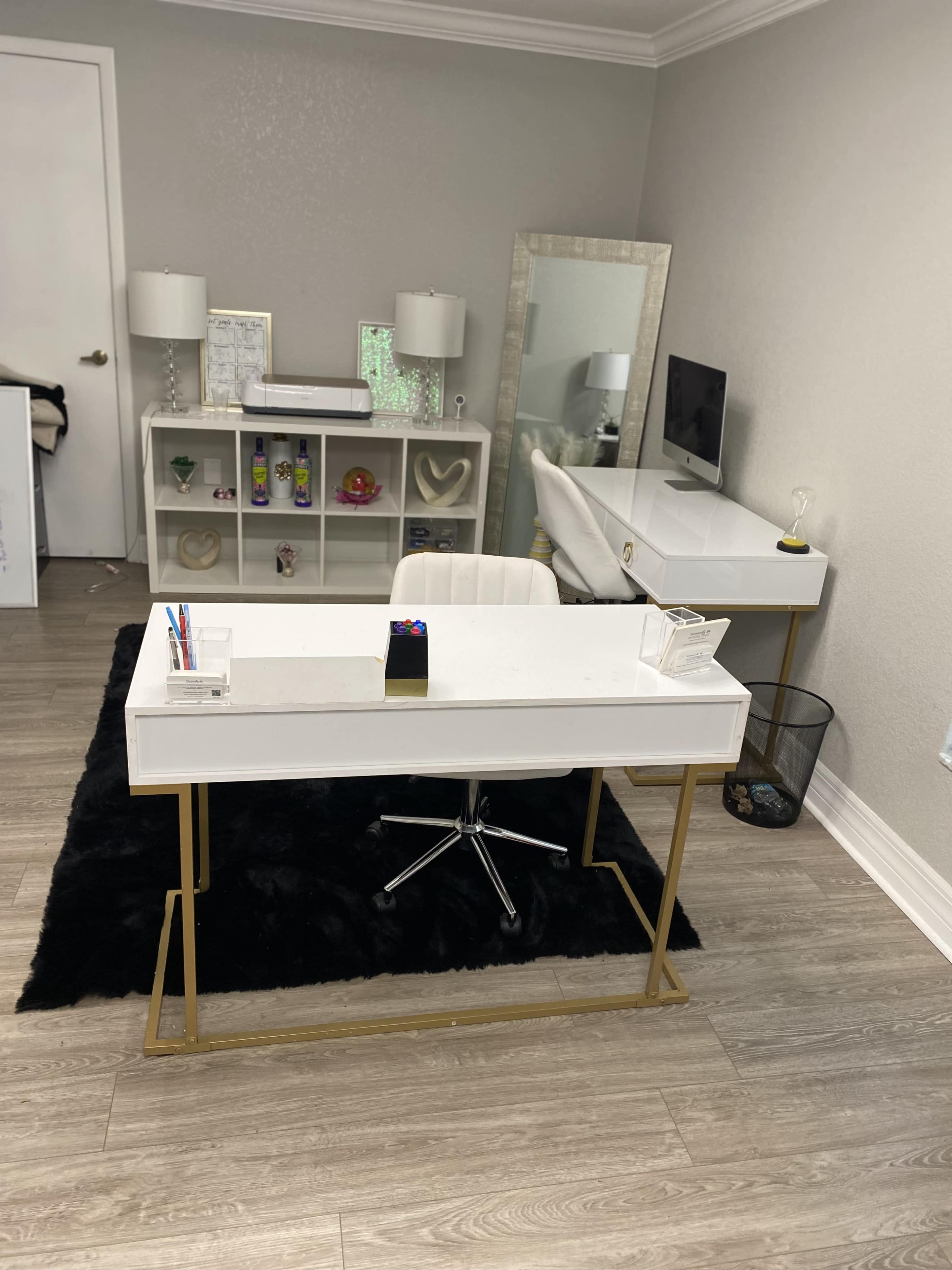 The image shows a modern office setup with a white desk on a black rug, accompanied by a chair, a shelving unit, a mirror, a lamp, and a wastebasket.