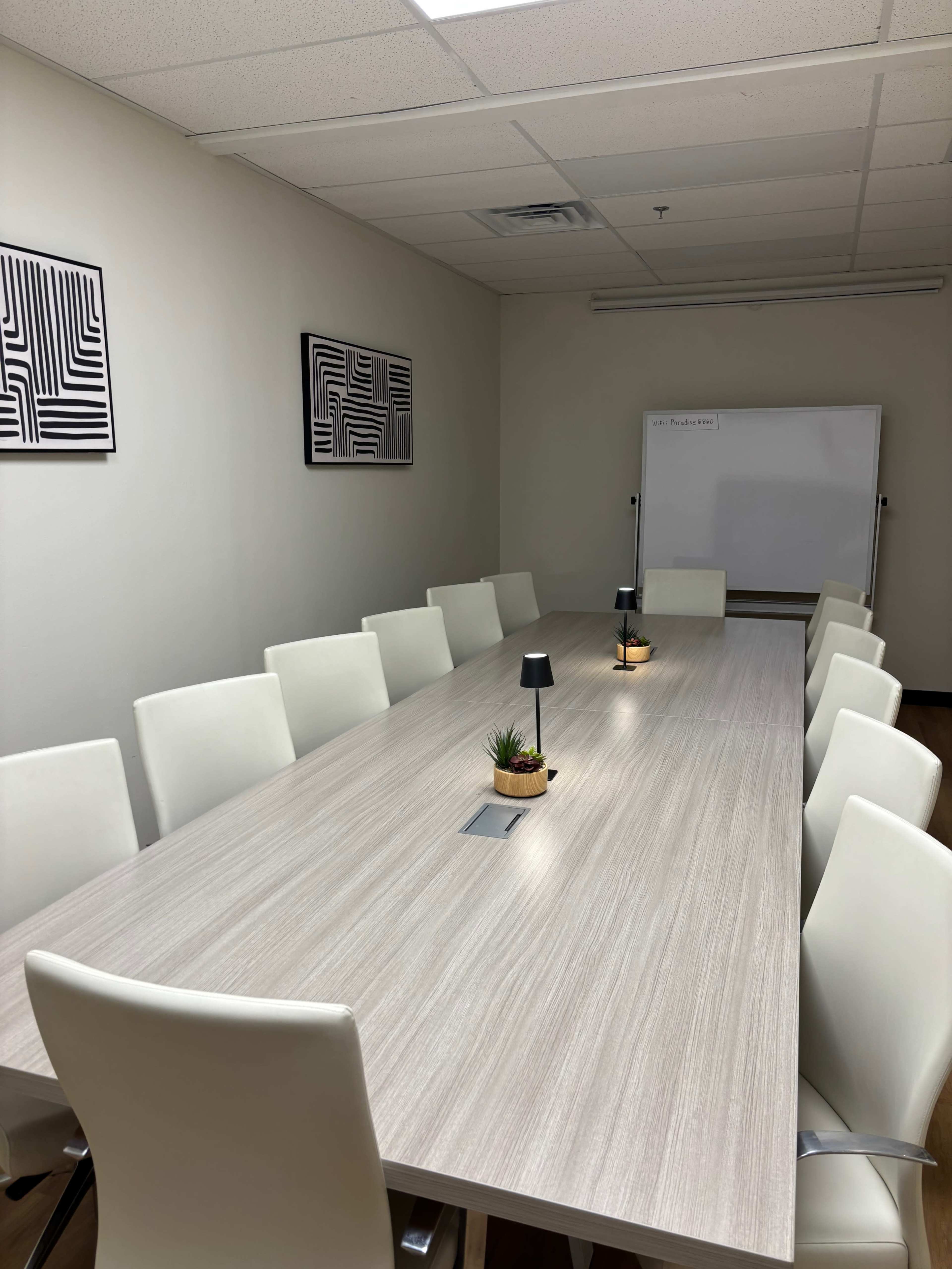 A long, rectangular conference table with white chairs is set up in a brightly lit meeting room, featuring two abstract wall art pieces and a whiteboard at one end.