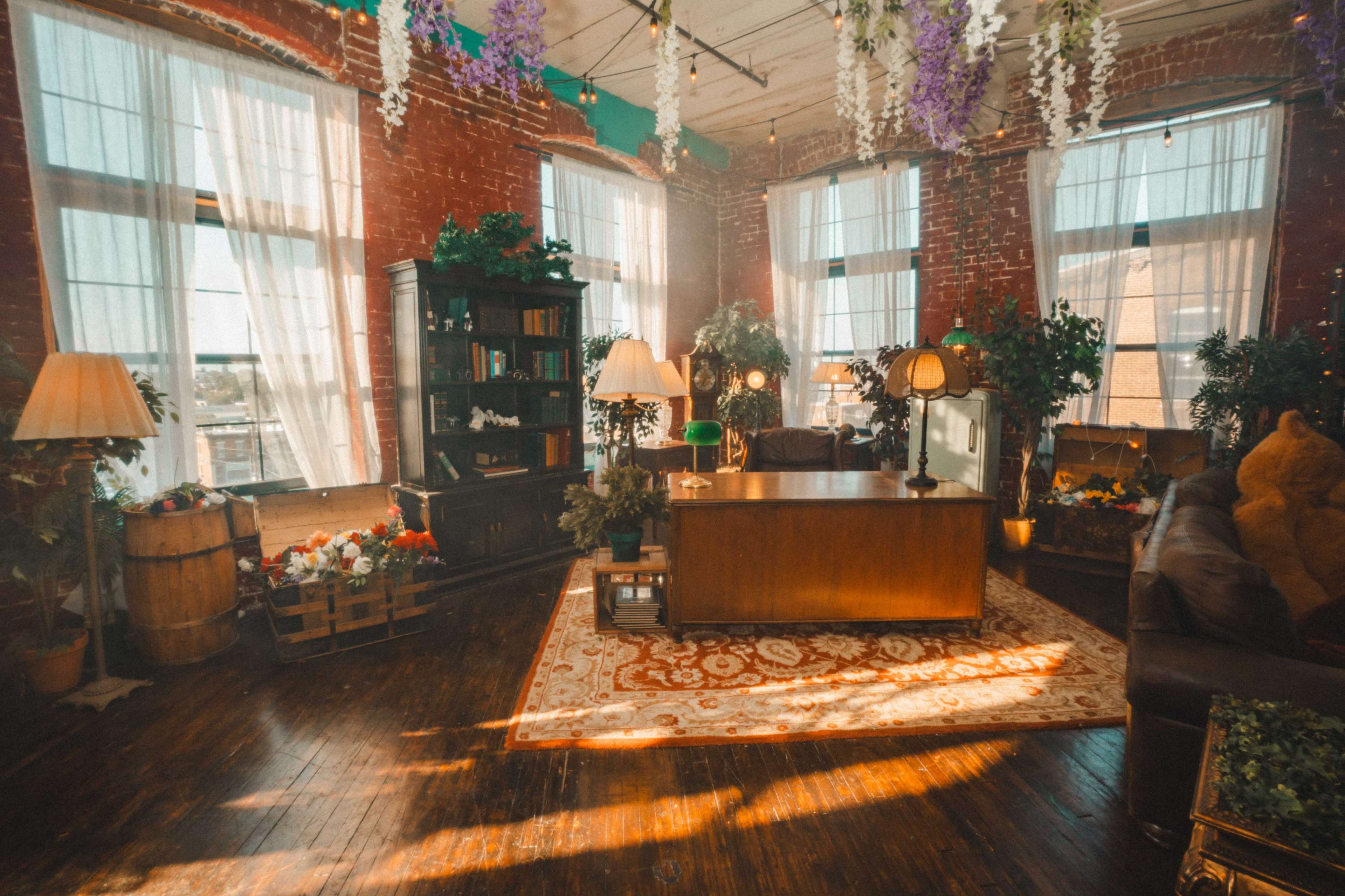 The scene features a cozy, warmly lit room with large windows, wooden flooring, and plants, complemented by a decorative rug and vintage furniture.