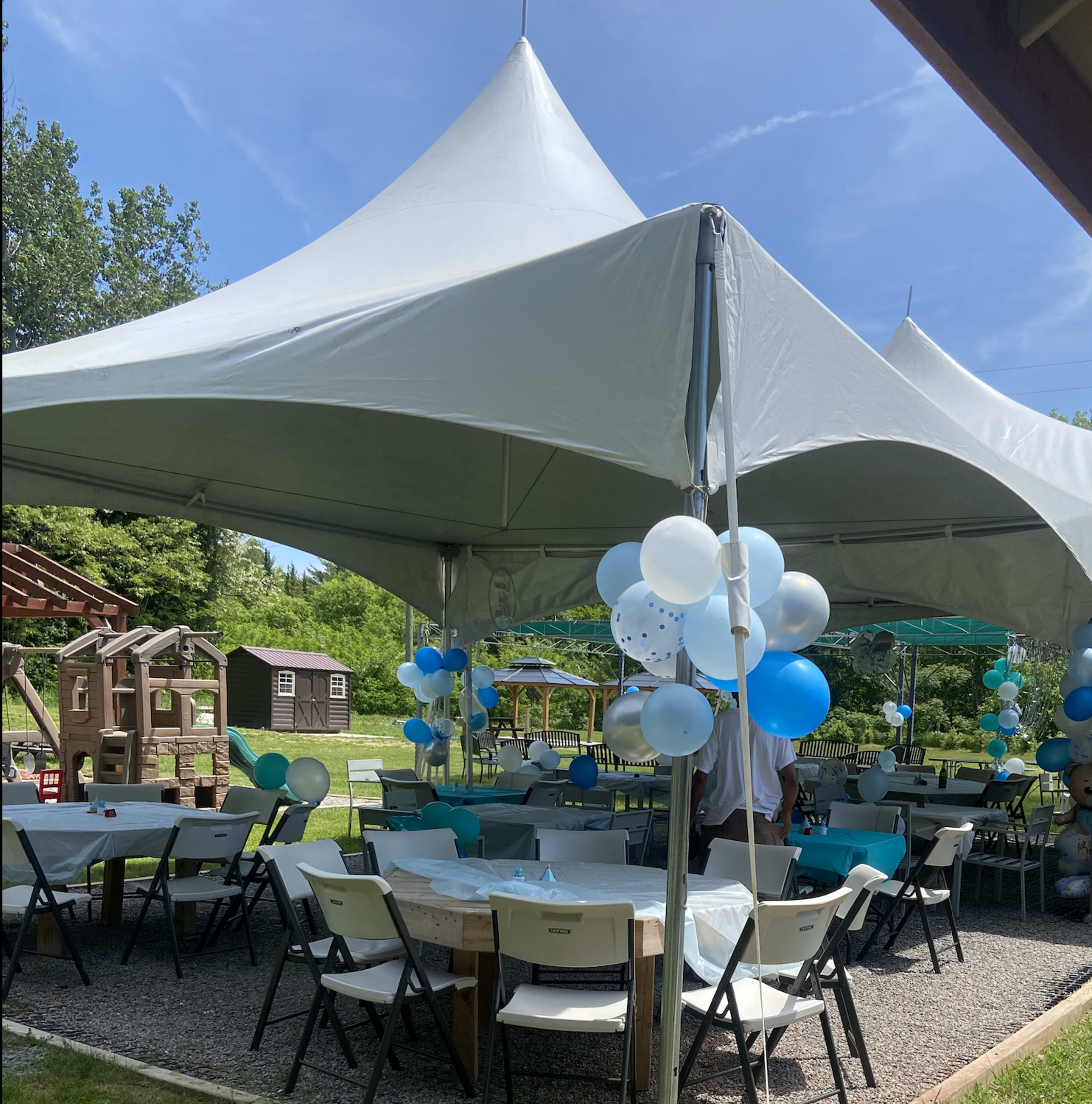 A large white tent is set up in an outdoor space, decorated with blue and white balloons, with several tables arranged underneath.