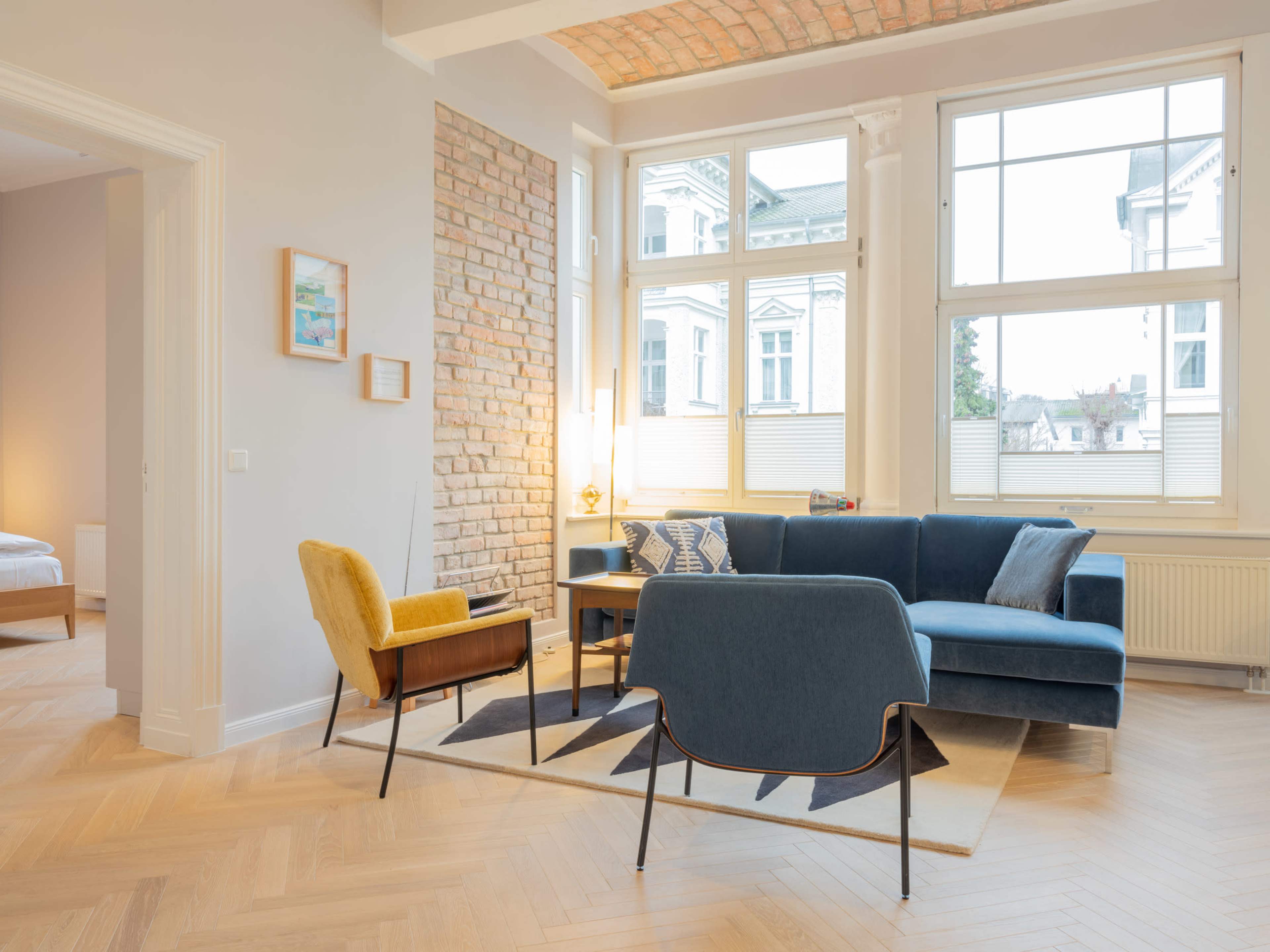 The image shows a modern living room with a blue sofa, yellow accent chair, and a large window allowing natural light to enter.
