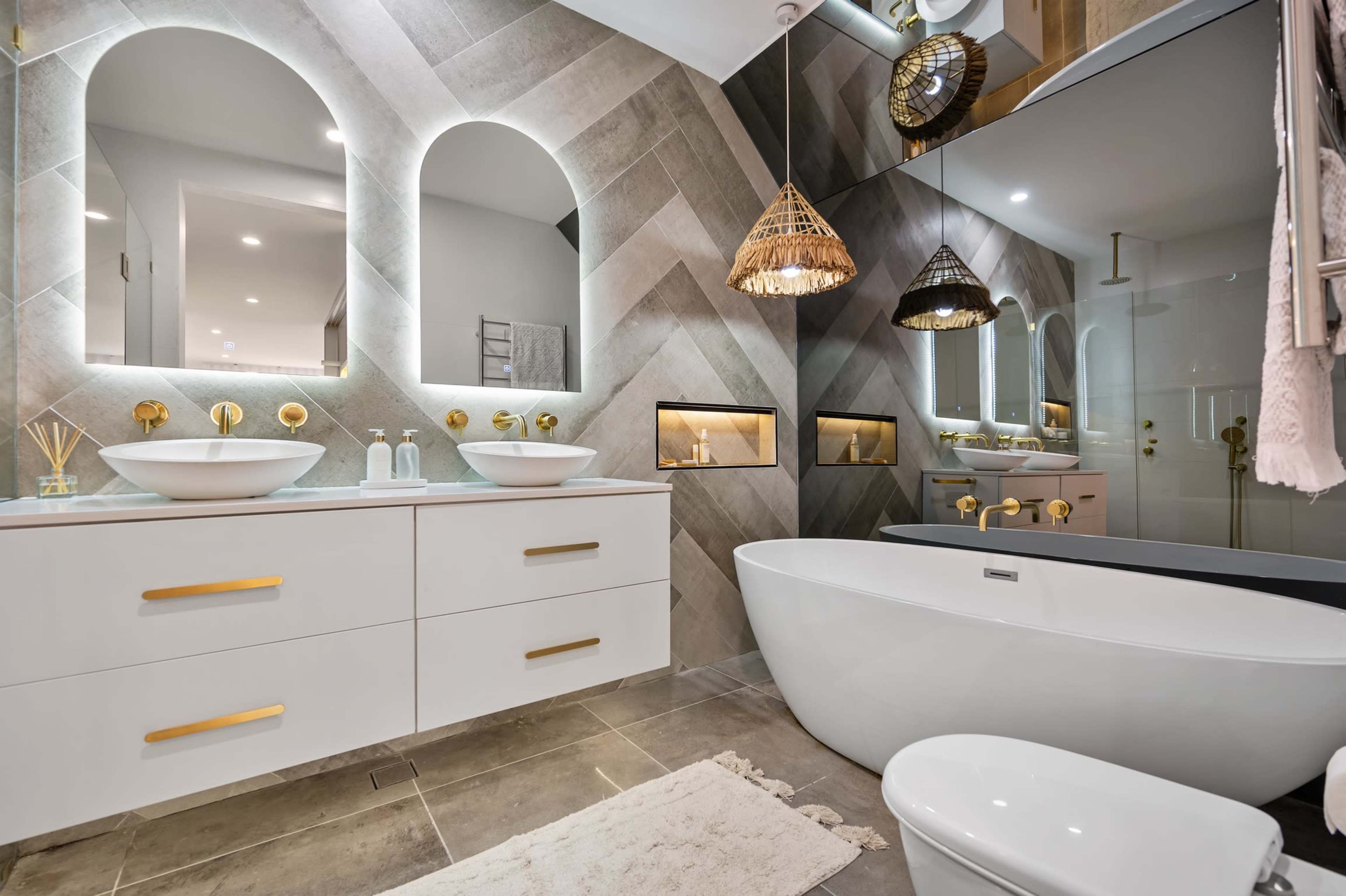 The bathroom features dual mirrors above a white vanity with gold fixtures, a freestanding tub, and textured tiled walls with built-in niches.
