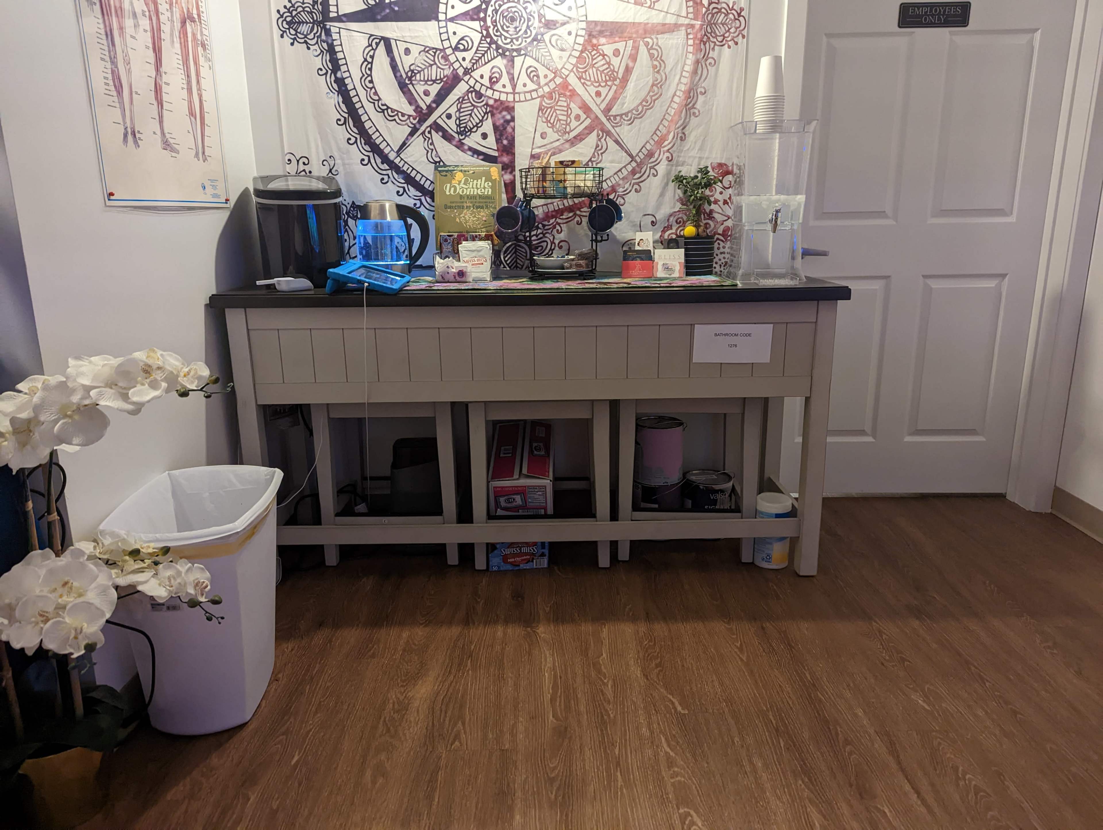 A beverage station with a counter, coffee machine, various drink supplies, and a trash bin, situated near a closed door and decorated with a large mandala.