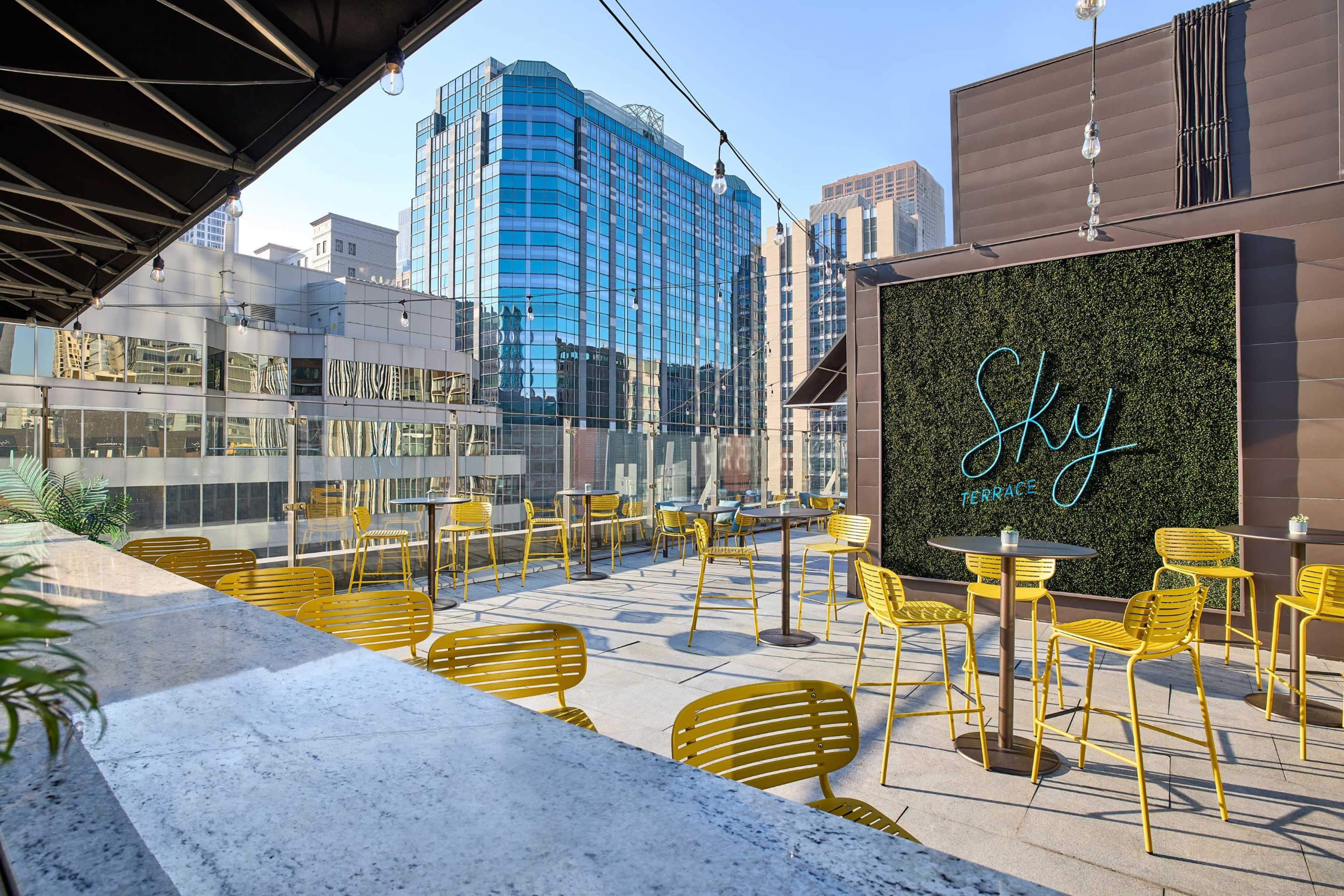 The image shows a rooftop terrace with yellow chairs and tables, surrounded by modern city buildings and a green wall featuring the word "Sky."