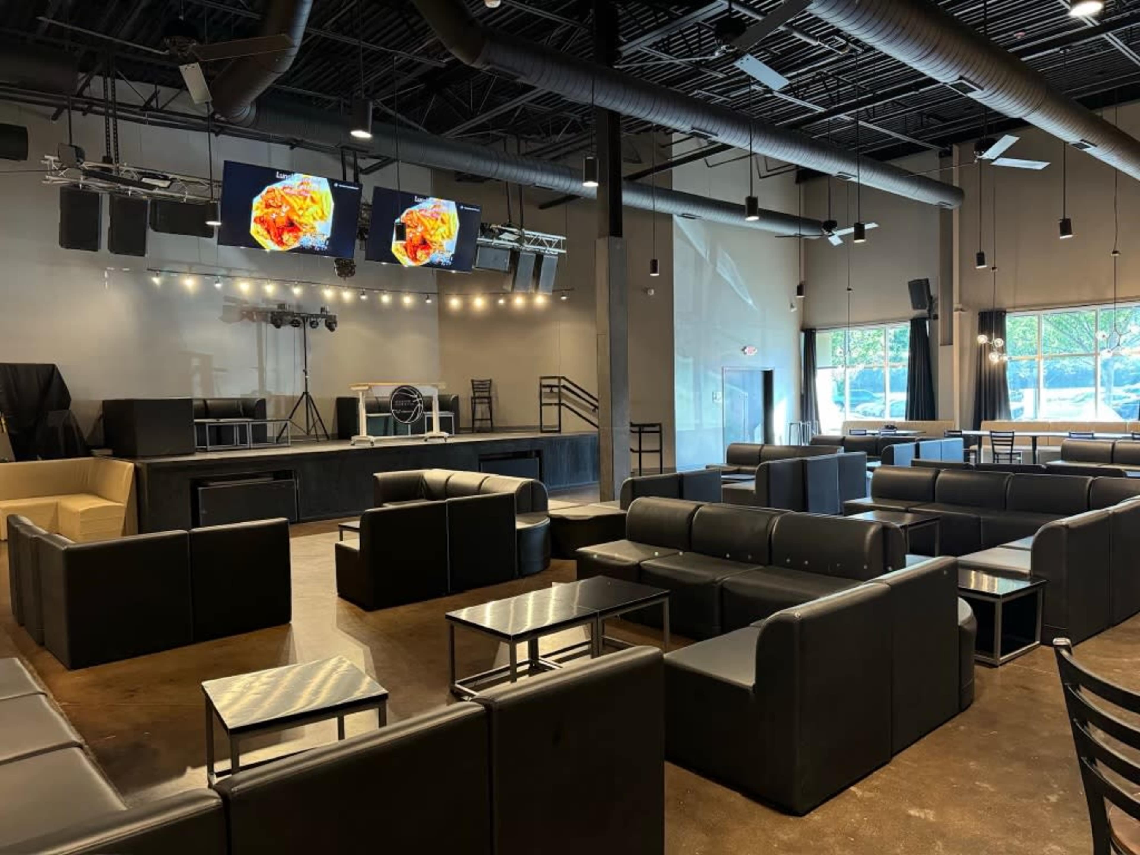 A spacious lounge area with black seating arrangements, a stage with lighting, and two large screens displaying images at the front of the room.