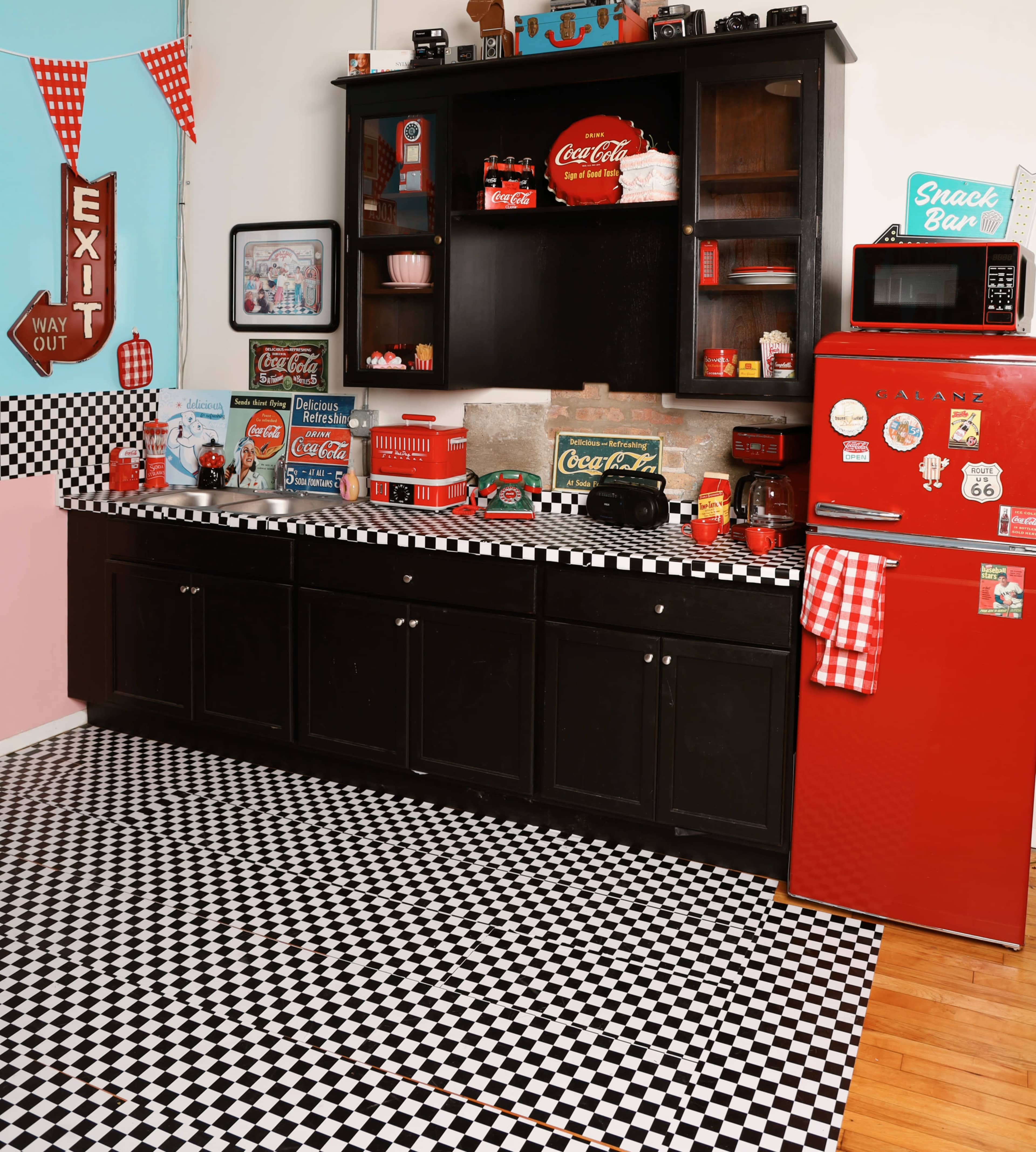 The image shows a retro-themed kitchen with black and white checkered flooring, a bright red refrigerator, and various Coca-Cola memorabilia displayed on shelves.