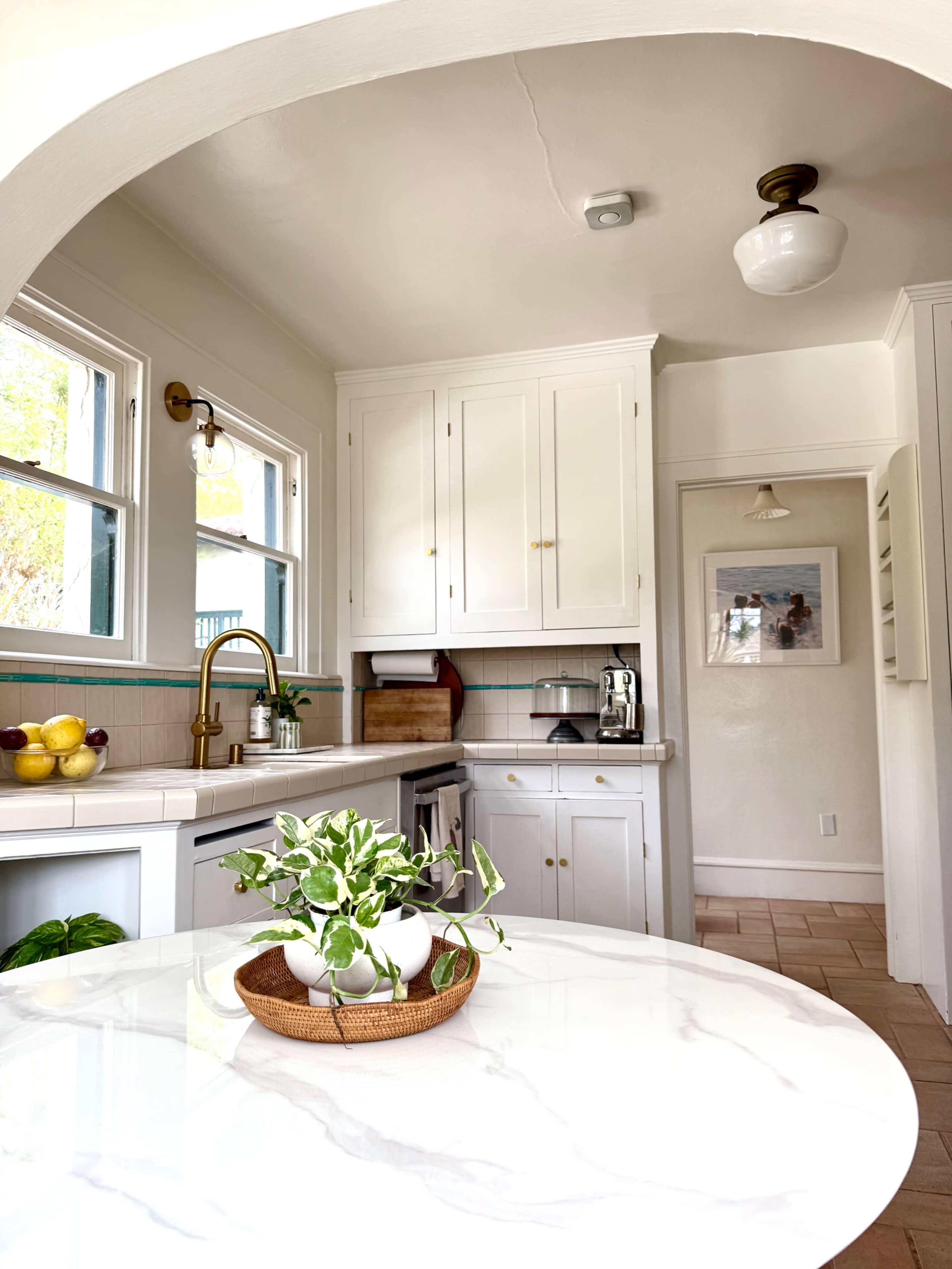 A bright kitchen features white cabinetry, a marble-topped table, and a small potted plant in the center.