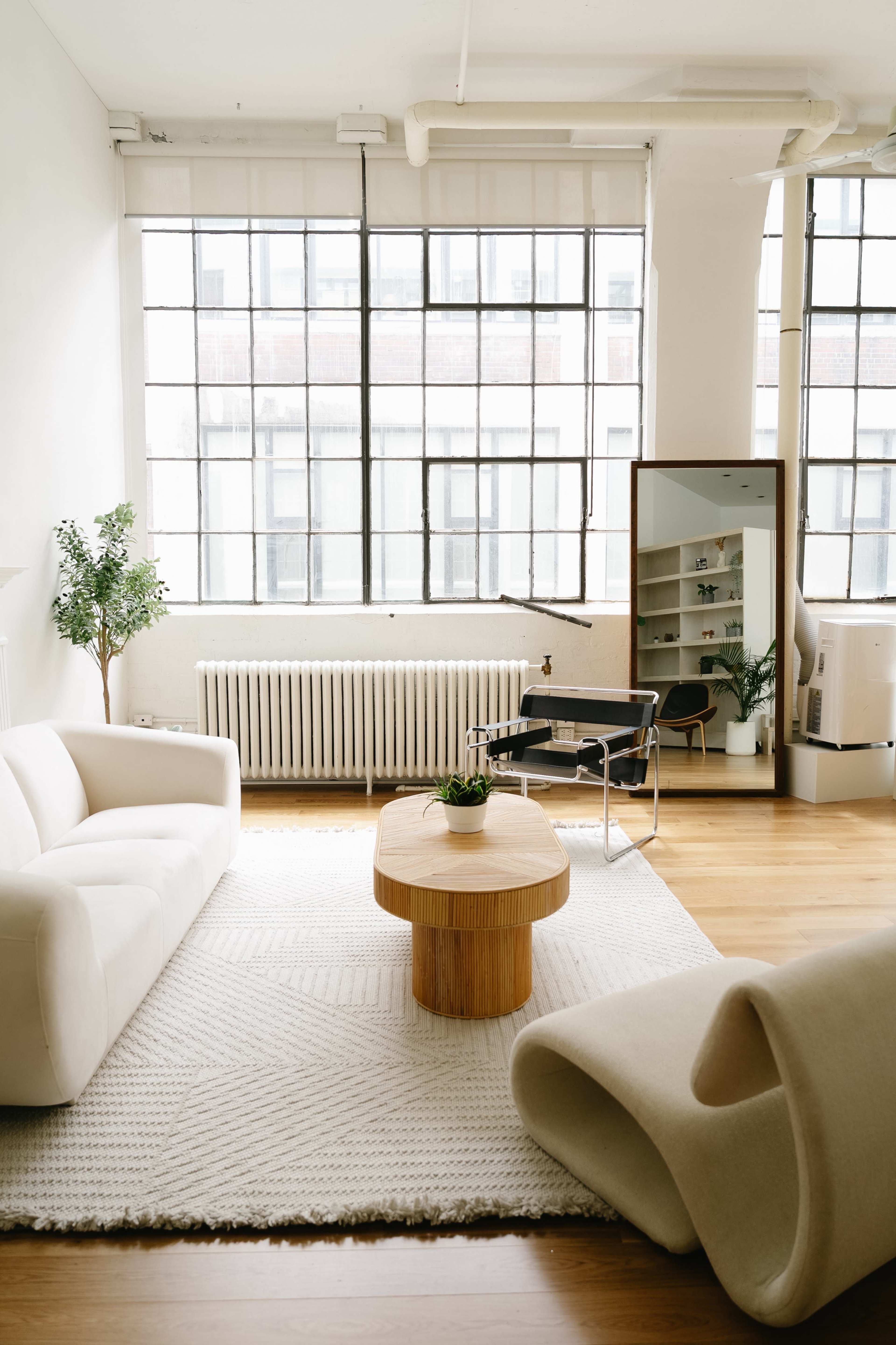 The image depicts a bright and airy living room featuring minimalist furniture, large windows, and a wooden coffee table at the center.
