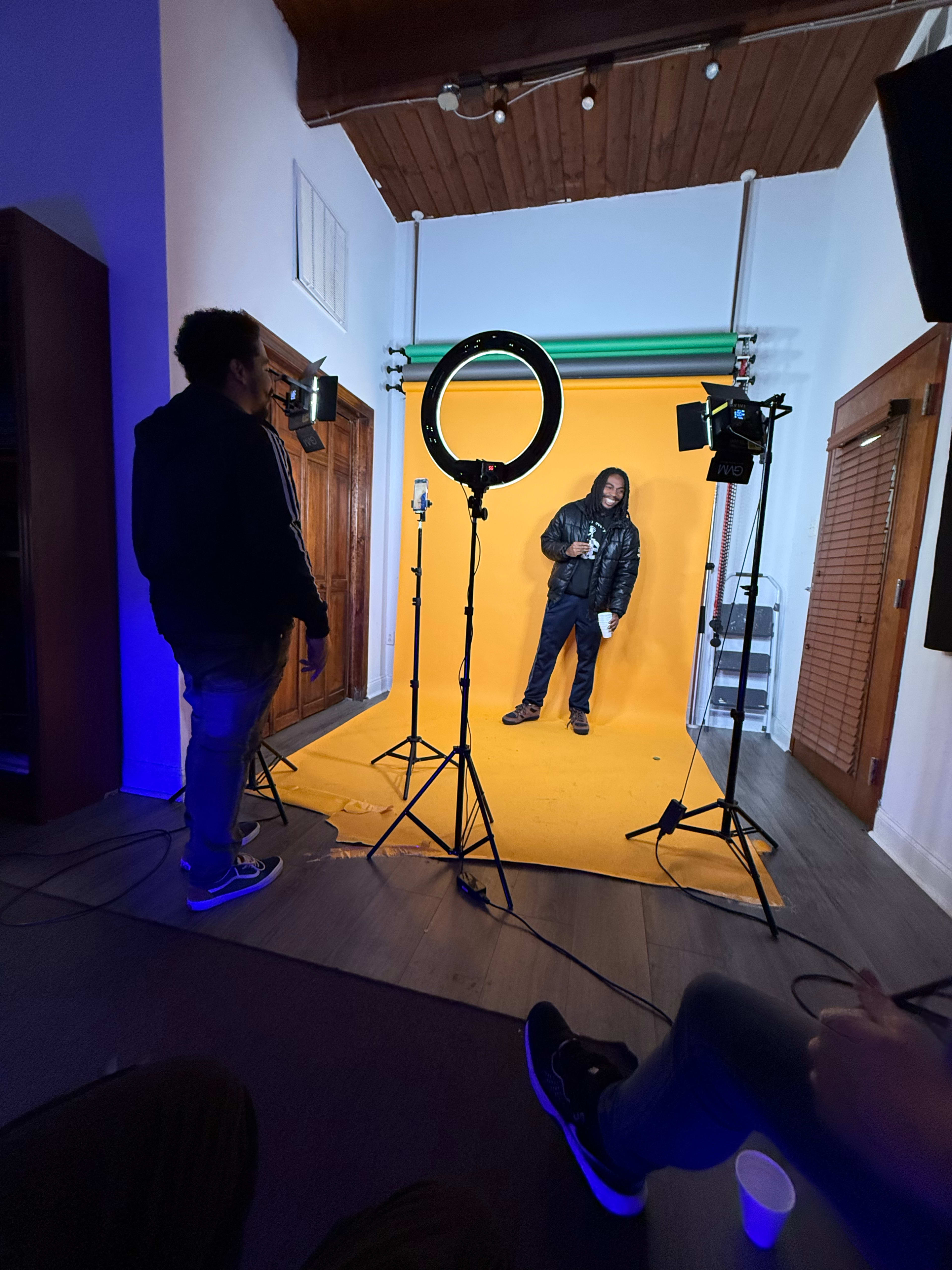 A person poses on a bright yellow backdrop in a photography studio with several lights set up around them.