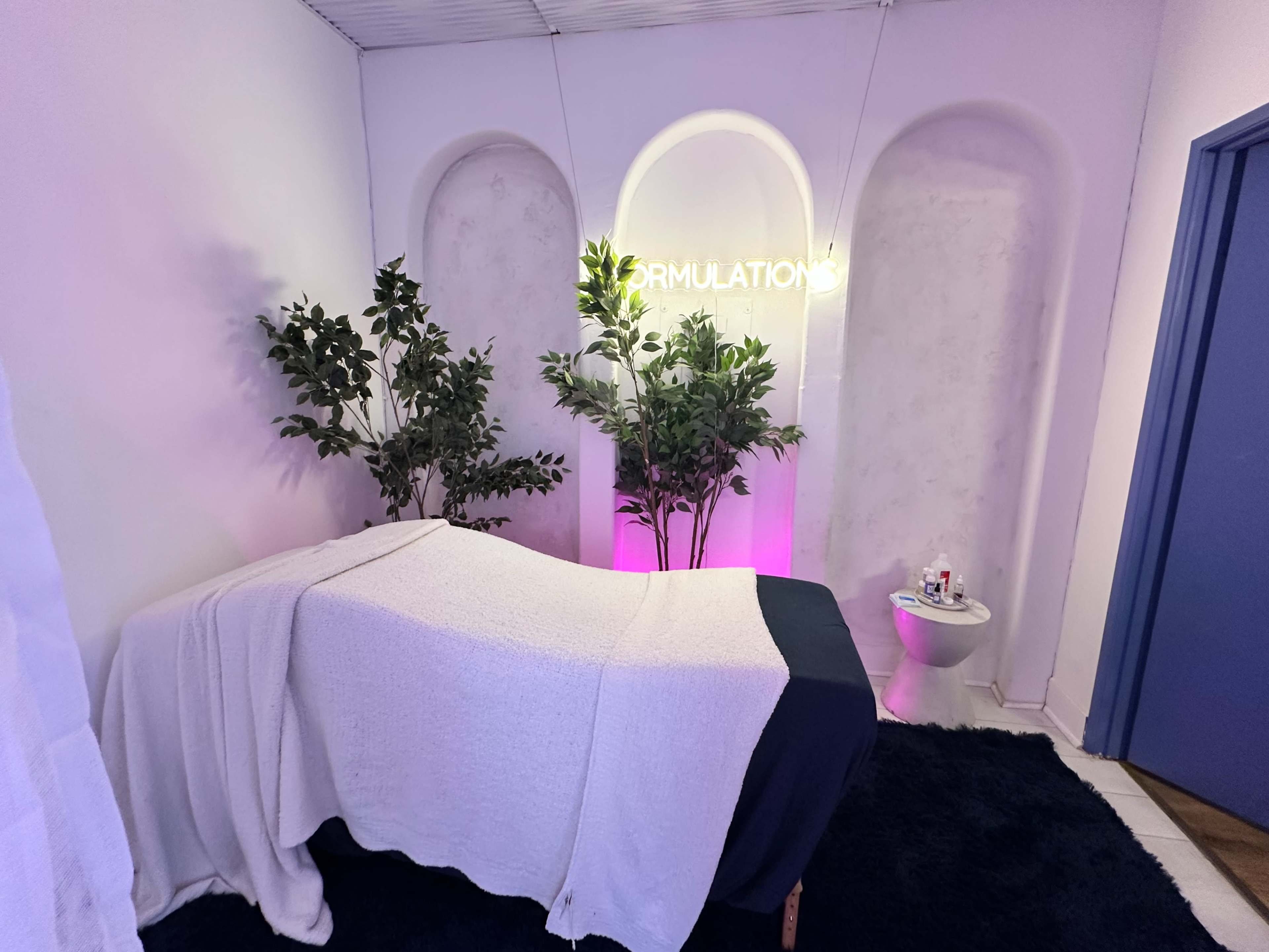 The image shows a serene treatment room with a massage table covered by a white sheet, two potted plants, and a neon sign that reads "FORMULATIONS" on the wall.